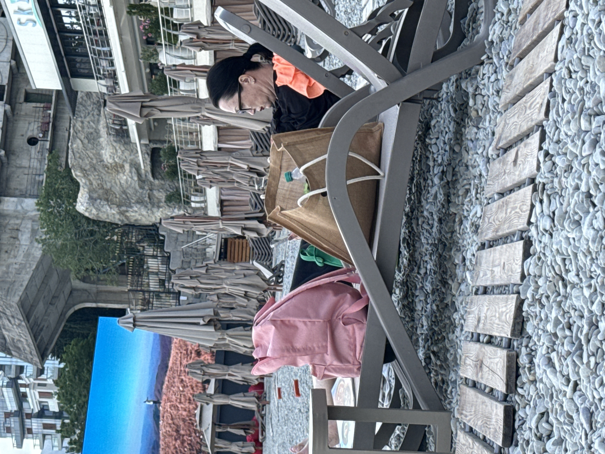 A woman with dark hair pulled back and glasses is seated in an outdoor lounge chair, looking down at a large brown tote bag. She is wearing a black shirt with an orange ruffled collar. The tote bag contains a plastic bottle with a green cap. Next to her, a pink backpack is visible. The scene appears to be a beach or outdoor seating area with many folded umbrellas in the background. Wooden planks are laid out on a bed of grey pebbles. In the distance, a large billboard displays a landscape with a blue sky and pinkish-brown terrain, featuring a small figure walking. The image suggests a relaxed daytime atmosphere. The location is identified as Yalta, Ukraine.