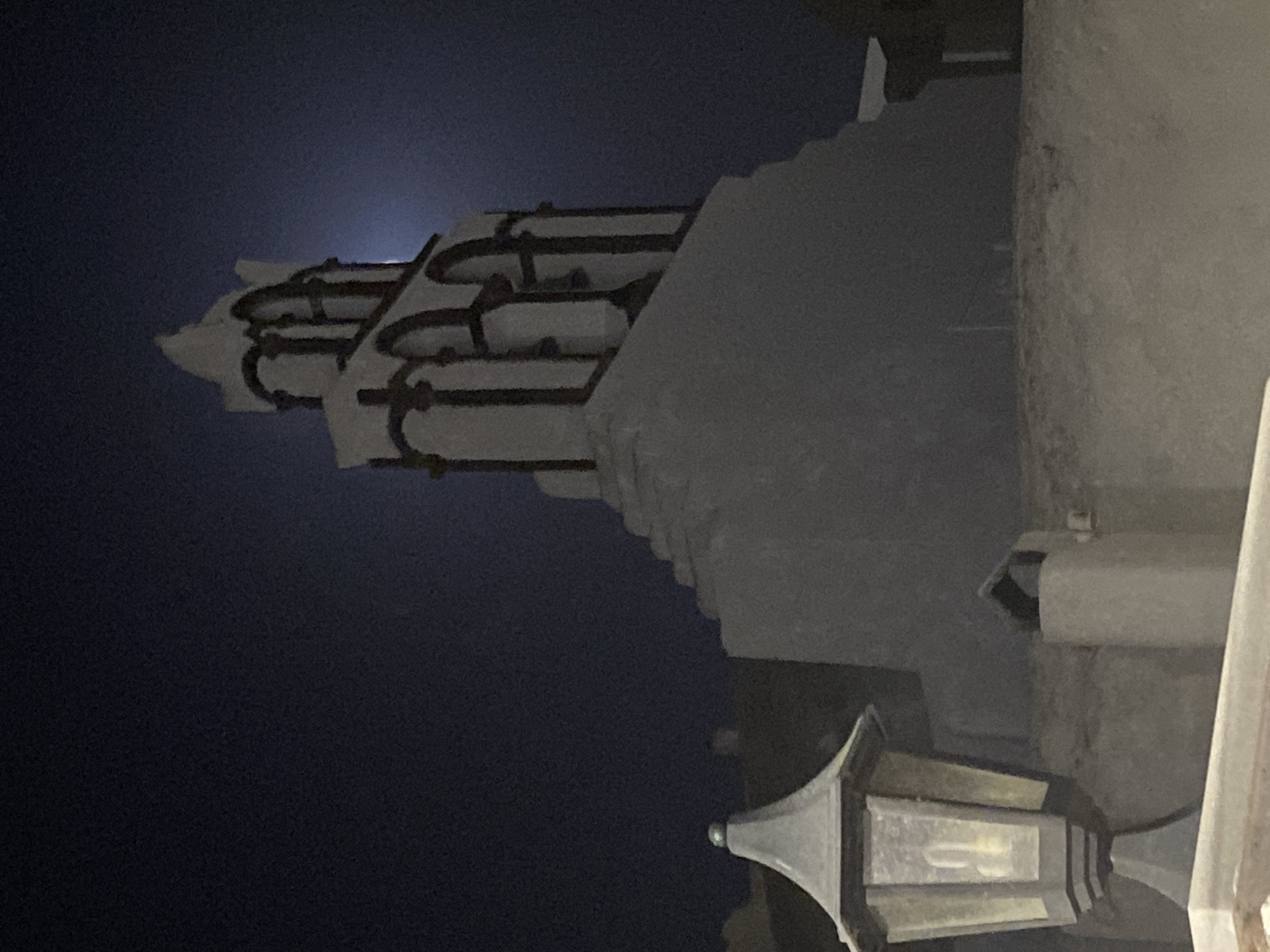 A night-time, low-angle, wide shot captures a white bell tower with dark arched openings against a deep blue, moonlit sky. The bell tower is situated in the city of Thira Municipal Unit, Greece, and appears to be part of a building complex with light-colored walls. A vintage-style lantern with a lit bulb stands in the foreground on the left, its glass panes illuminated. The overall scene evokes a sense of tranquility and historical charm under the moon's glow.