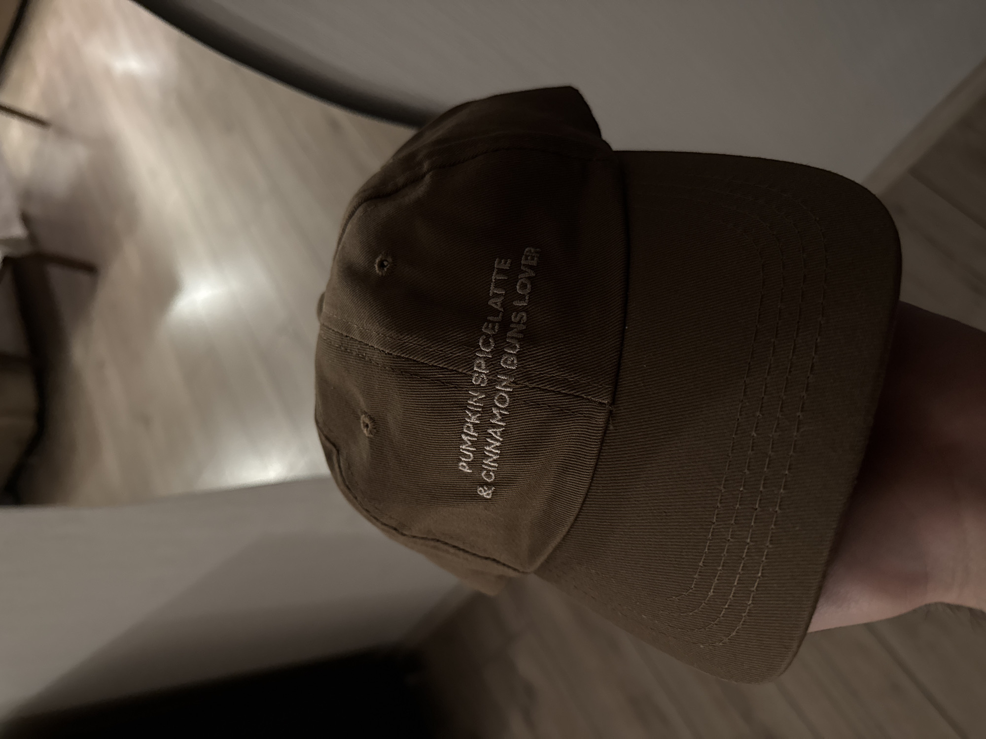 A brown baseball cap is held in a hand, with the brim facing the viewer. Embroidered on the cap in a lighter brown thread are the words "PUMPKIN SPICELATTE & CINNAMON BUNS LOVER". The background is blurred and appears to be an indoor setting with wooden flooring.