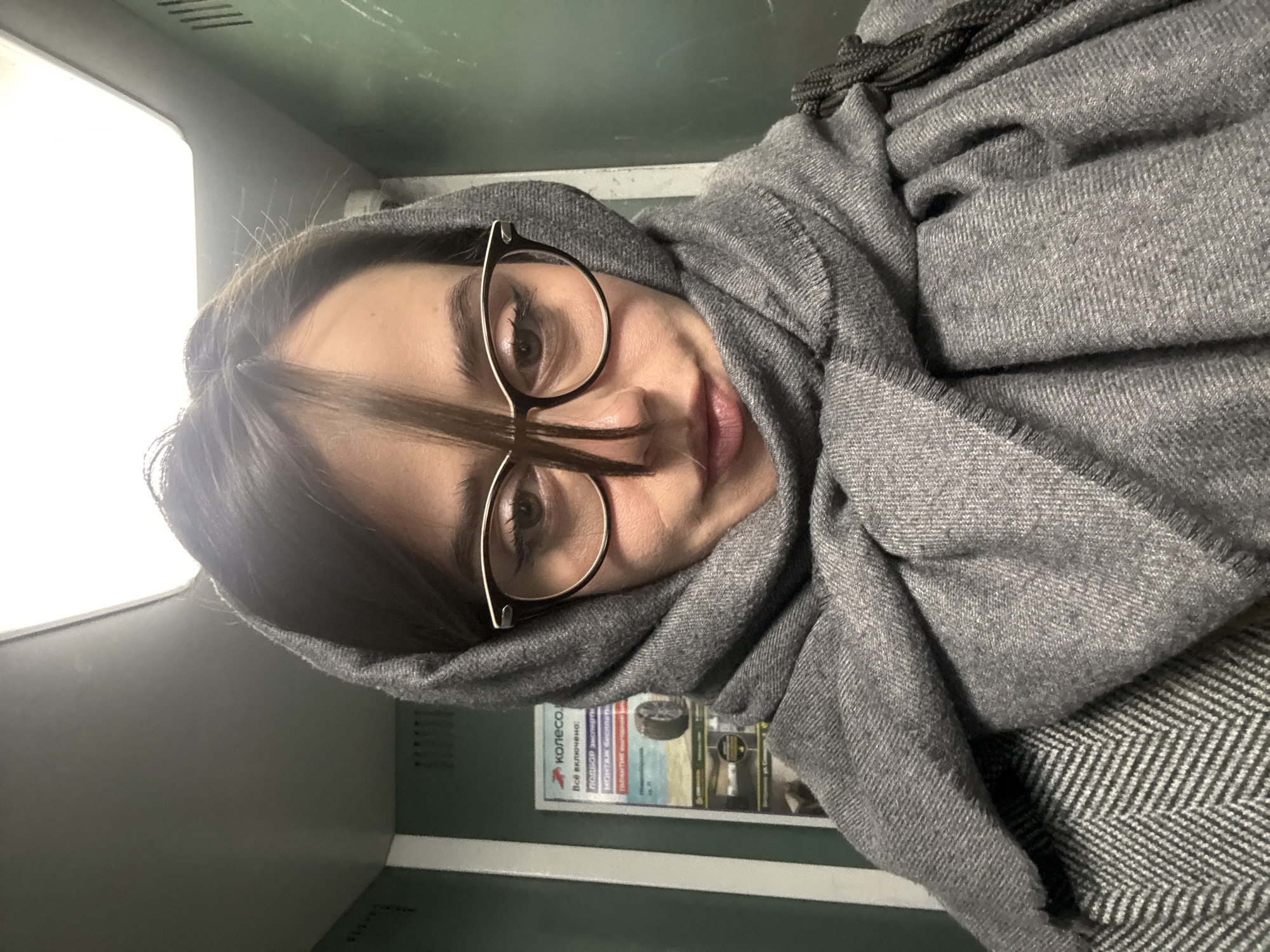 A woman with brown hair and glasses is sitting in what appears to be a public transportation vehicle, possibly a train. She is wearing a gray scarf wrapped around her head and shoulders. Behind her, a promotional poster advertises "KОЛЕСО" (Koleso), which is "wheel" in Russian. The poster contains a picture of tires and mentions services like tire selection, installation, balancing, and repairs. The poster indicates that this is likely located in Krasnoyarsk, Russia, as the poster is in Russian and Krasnoyarsk is a major city in Russia. The overall lighting seems to be dim.