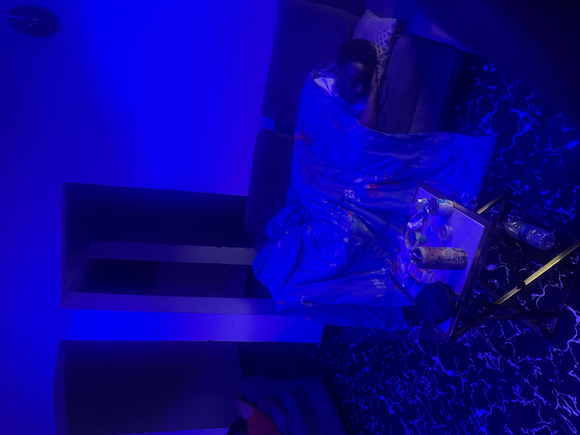 The image depicts an indoor scene, likely in Abuja, Nigeria, characterized by strong blue ambient lighting that casts a dark, moody atmosphere. A person, appearing to be an adult, is seen resting or sleeping on a dark-colored sofa or bed, covered by a blue blanket adorned with a subtle floral or abstract pattern. The person's face is partially visible, indicating a state of repose.
Beside the person, a stylish side table with a reflective top and metallic frame holds several items. These include at least two plastic water bottles, one opaque bottle or can with partially visible text "HAR..." or "HAP...", and other small, indistinct objects. The floor beneath the table features a distinctive black and white or black and blue marble-like pattern. To the left, a dark wall or shelving unit is also bathed in the pervasive blue light. The intense blue illumination suggests a nighttime setting or a deliberate lighting choice, creating a serene and somewhat futuristic ambiance.