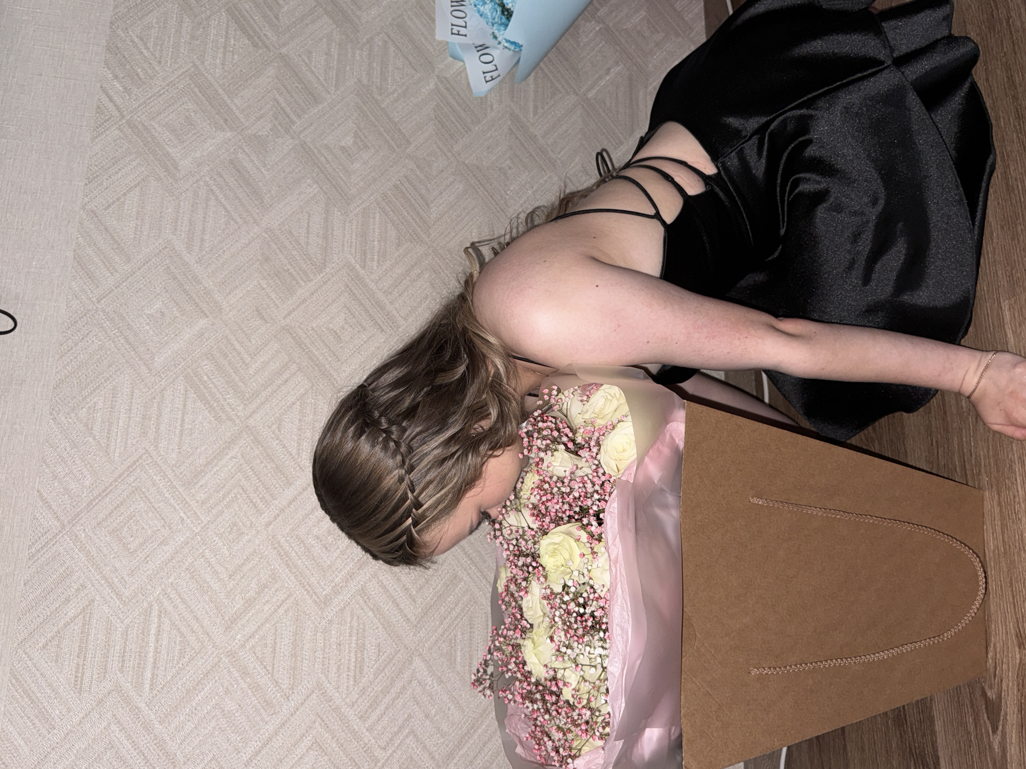 A young woman is lying on a patterned carpet, her body angled towards a large bouquet of white roses and pink filler flowers presented in a pink tissue paper within a brown paper shopping bag. She wears a black satin dress with thin straps and a criss-cross design on the back. Her hair, styled in a braid and left to fall over her shoulder, partially obscures her face as she leans into the flowers. Her skin appears flushed. A light blue gift box with "FLOW" written in white text twice on a ribbon is visible in the background, lying on the carpet. The lighting suggests an indoor setting, possibly a home. The overall mood conveyed is one of quiet appreciation or joy.