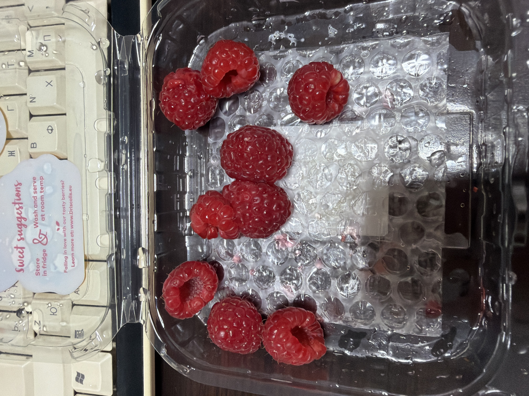 A close-up shot shows a clear plastic container of raspberries sitting on a keyboard. The keyboard appears to be a vintage model with off-white keys and a clear plastic cover, and is to the left of the container. A small sticker is visible on the keyboard cover with text "Sweet suggestions, store in fridge & wash and serve at room temp, Falling in love with our tasty berries? Learn more at: www.Driscolls.eu". The raspberries are arranged on top of a piece of bubble wrap inside the container. Some water droplets are visible on the plastic surfaces. The background appears to be a dark, wooden surface. The scene appears to be indoors. There is no location in Pleven, Bulgaria, to identify, nor are there any people, actions or interactions to describe in this scene.