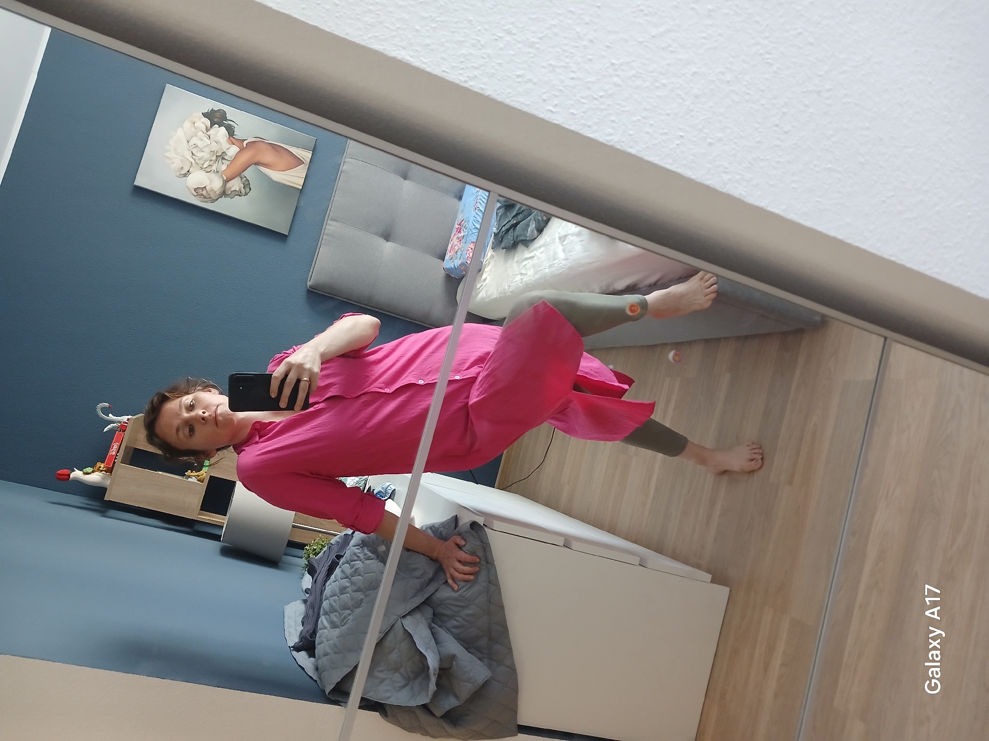 A woman in a bright pink dress and olive green leggings is taking a selfie in a mirror. The reflection shows a bedroom. Visible are a bed with a grey headboard, a blue accent wall with a floral artwork, a white dresser with a grey comforter on top, and a wooden floor. The "Galaxy A17" text is visible in the lower right corner of the reflection, indicating the phone model used for the selfie. The location of Yekaterinburg, Russia, cannot be determined from the visual information provided.