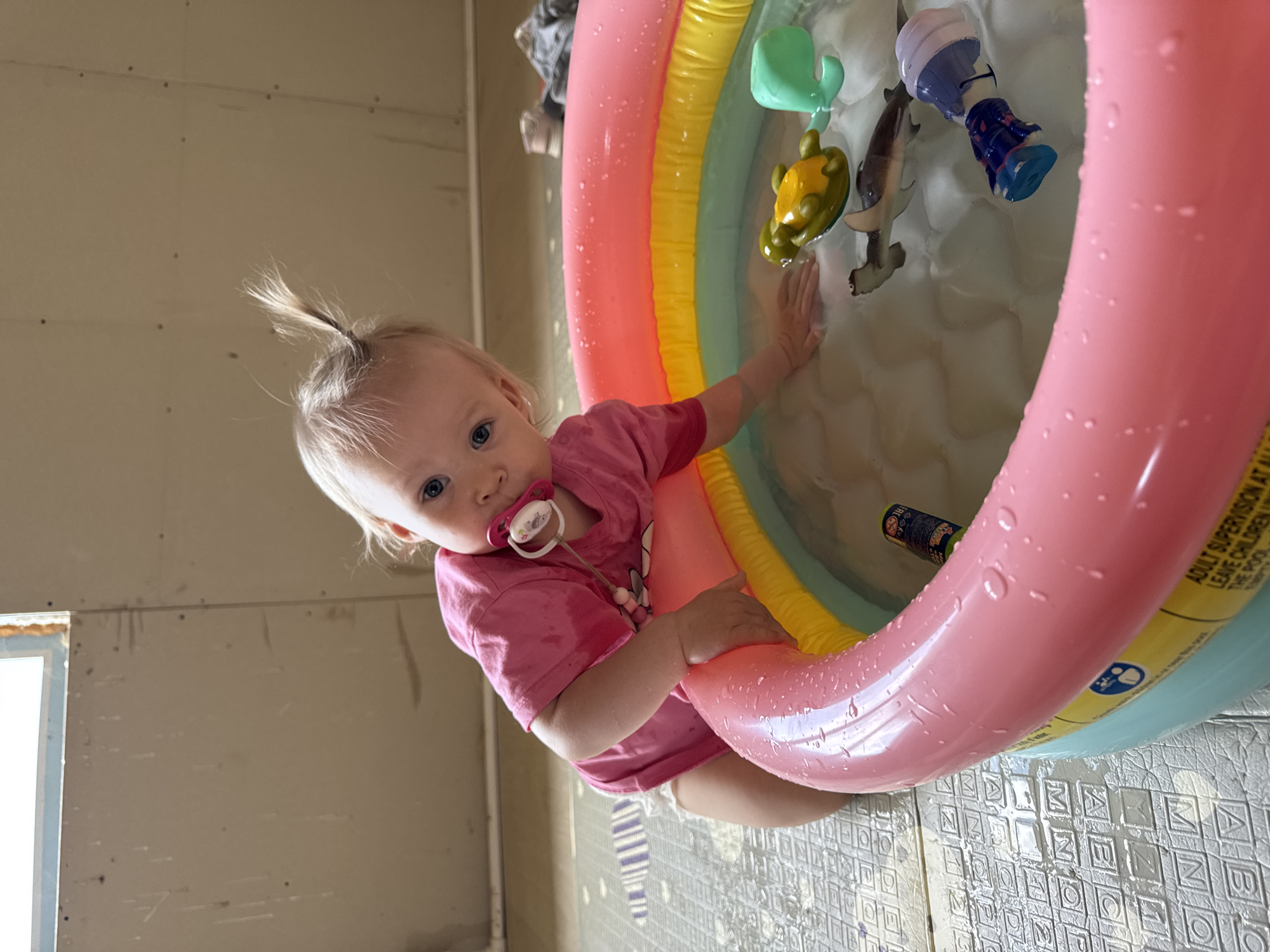 A baby wearing a pink shirt and a pacifier is reaching into a small inflatable pool filled with water and toys. The toys include a green whale, a yellow turtle, a hammerhead shark, a blue and white boot-shaped toy, and a dark cylindrical object. The pool is pink, yellow, and green. The room has unfinished walls and patterned flooring. The location is Фоки, Russia.
