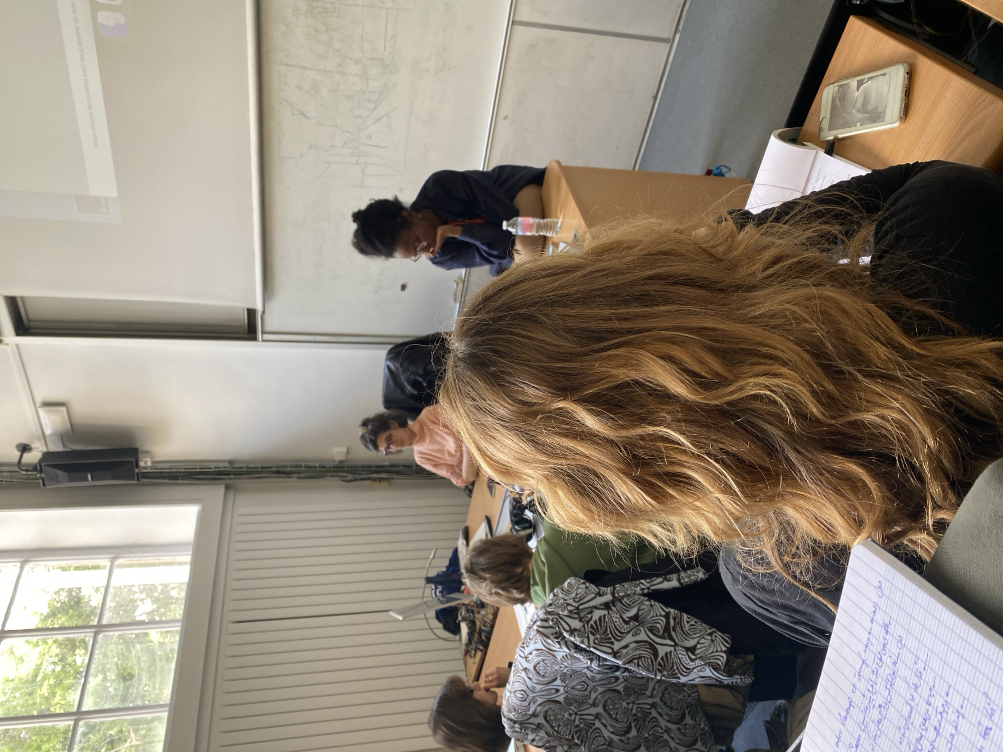 A classroom scene in Paris, France is depicted. Several students are seated at desks. At least six individuals are visible, each engaged in activities associated with classroom learning.
One student, identifiable by their dark hair pulled back in a bun, is wearing a navy blue garment and appears to be focusing on something on their desk. A water bottle is placed near them on the desk surface.
In the foreground, a person with wavy blonde/brown hair sits with a notebook in front of them, possibly taking notes. A student in a patterned shirt can be seen in the middleground. Additional students are further back, including one wearing a pink shirt.
The environment is a typical classroom, with desks, chairs, and a whiteboard mounted to the wall. Light enters the room through a window, suggesting it's daytime. The desks are arranged in rows. There's also a mobile phone lying on one of the desks.