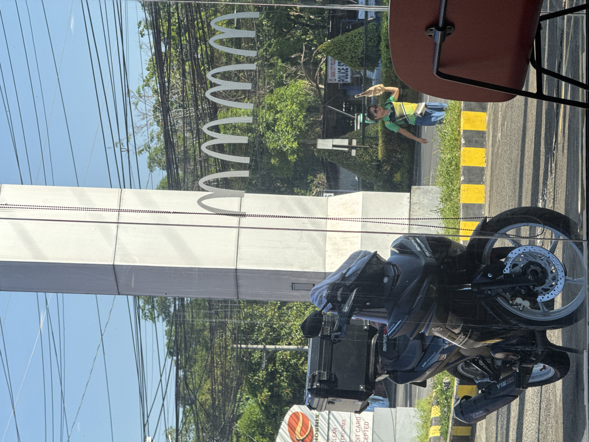 Captured from inside a building, likely a fast-food restaurant, this image shows a bright, sunny street scene in Davao City, Philippines, viewed through a glass window. The glass features the distinctive white arches of a McDonald's logo, partially visible as "wwww" due to the perspective and reflection.
In the foreground, inside the building, a portion of a red chair and table is visible. The window itself reflects the vibrant outdoor environment.
Outside, a man is the primary human subject, seen standing or walking on a sidewalk. He is wearing a green and yellow striped polo shirt, blue pants, and sunglasses, holding a light-colored bag or object in his right hand. Beside him, a black and yellow striped curb separates the sidewalk from the road. Further back, numerous trees and overhead power lines are reflected in the window.
A black Yamaha Sniper 155 motorcycle is prominently parked outside, reflecting against the glass. It features a large black cargo box on the rear. Specific text visible on the motorcycle includes "SNIPER 155", "VVA", and "YIEZAR".
Further into the background, reflections reveal signs for "DAVAO CITY POLICE" and "POLICE ST", confirming the location. Inside the building, near the bottom edge, parts of a "PHOENIX" logo, "CLEAN RESTROOMS", and "CREDIT CARD ACCEPTED" signs are also visible through the window's lower section. The bright daylight suggests it is daytime.