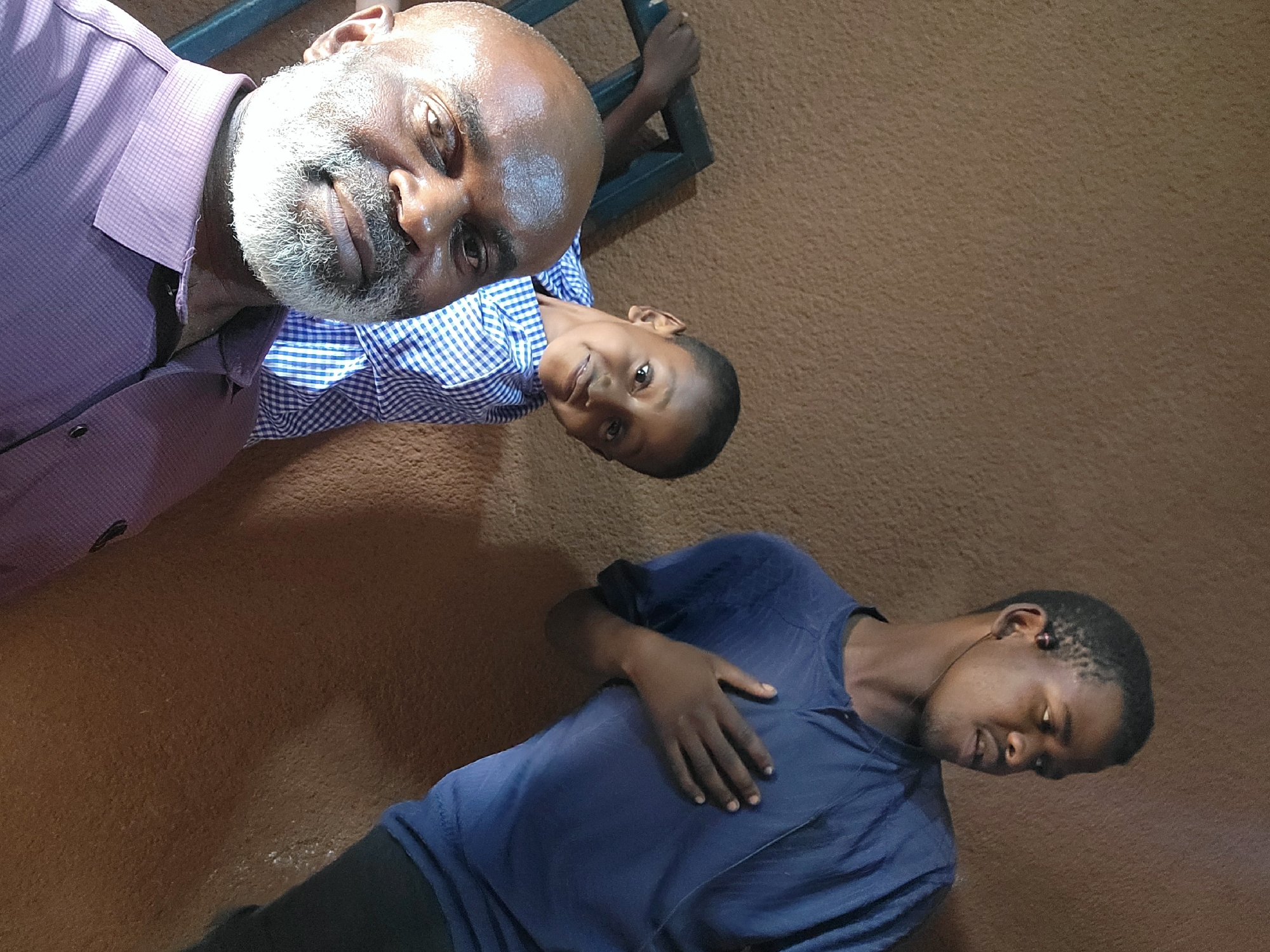 The image is a casual portrait featuring three individuals of African descent, set against a consistent, textured earthy brown background, suggesting an indoor space or a sheltered outdoor area. The photograph is oriented such that the subjects are arranged diagonally from the upper left to the lower right.
In the upper left portion of the frame, an older man with dark skin, a bald head, and a prominent white/grey beard is visible. He wears a light purple checkered collared shirt and looks directly at the camera with a gentle, friendly smile.
Adjacent to him, positioned slightly below and to the right, is a young boy with dark skin and short dark hair, wearing a blue and white checkered shirt. He also smiles faintly while looking towards the camera. His right hand is seen grasping a dark, possibly metallic, structure or railing behind him.
Further down and towards the lower right, a younger man with dark skin and short dark hair is depicted. He is dressed in a dark blue t-shirt and dark pants. His gaze is directed downwards or away from the camera, and his left hand rests on his chest. Small earbuds or headphones are visible in his ears.
The overall atmosphere appears relaxed and informal, suggesting a casual gathering or a posed photograph. There is no specific activity taking place beyond the subjects' presence. The lighting is uniform and soft, preventing clear determination of the time of day or outdoor conditions. There is no visible text within the image. The provided context indicates the location is Maiburiji, Nigeria.