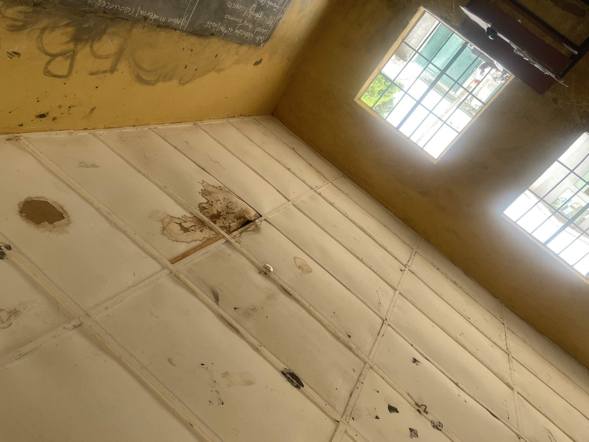 The media file captures an upward-looking view of the interior of a dilapidated room, likely a classroom, in Unguwar Rimi, Nigeria. The scene is dominated by a white ceiling composed of square panels, many of which show significant water damage, staining, peeling paint, and missing sections, revealing the wooden framework beneath. Several panels have large brown water stains, and at least two areas show large holes where panels have fallen away.
The walls, painted a faded and dirty yellow, meet the ceiling. On one wall, partially visible, is a blackboard with educational content written in white chalk. The visible text includes "Date: Wednesday 22nd April 2026," "Subject: Home Economics," "Topic: Family Resources," and "Non material resources." Below the blackboard, on the yellow wall, large, faded graffiti spelling "BB" is visible.
Two rectangular windows with metal grilles are cut into the upper part of the wall, letting in bright daylight. The light illuminates the dust and grime on the window frames and casts a strong glow, suggesting it is daytime. Through the windows, glimpses of green foliage from trees and what appears to be another building or structure are visible, hinting at an outdoor environment. Above one window, a dark-colored shelf or piece of furniture is mounted. The overall condition of the room, with its damaged ceiling, dirty walls, and simple furnishings, indicates a setting that is poorly maintained or in disrepair. There are no people visible, and no direct activity is taking place.