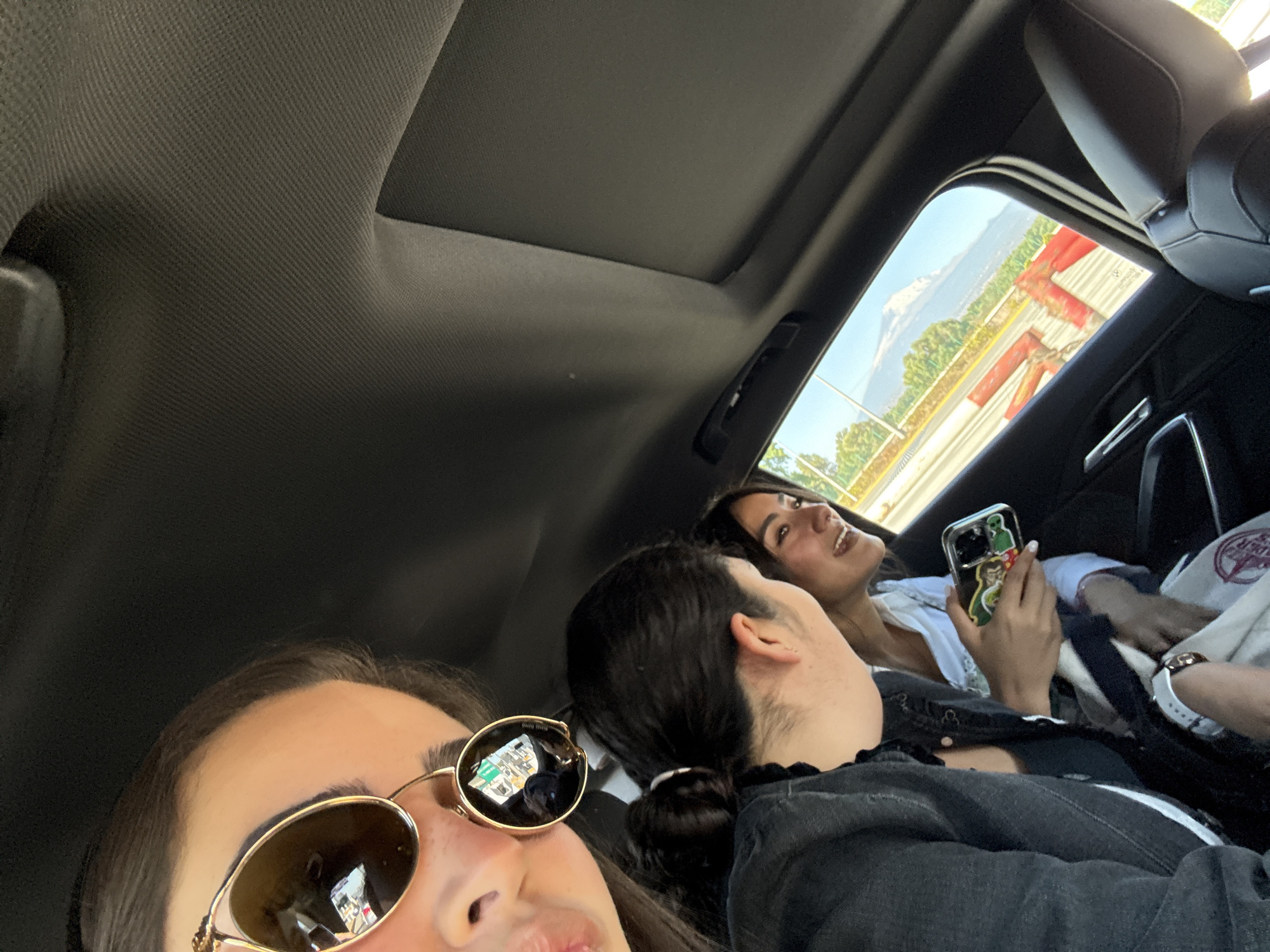 The image captures the interior of a vehicle with three occupants, situated in or near Santa María Moyotzingo, Mexico, based on the external landscape.
In the immediate foreground, the upper left portion of a person's face is visible, wearing large, round, gold-rimmed sunglasses with dark lenses. Reflections in the sunglass lenses show ambient light and indistinct interior details.
To the right of the foreground person, a second individual is seen from a side and rear perspective. This person has dark hair pulled back into a bun or ponytail and wears a dark upper garment. A white-banded watch is visible on their left wrist. They are turned towards a third individual, suggesting interaction.
The third individual is positioned further back on the passenger side. This person has dark hair, wears a light-colored top, and is smiling while looking at the second individual. They hold a smartphone with a colorful, character-themed case, specifically identifiable as Mickey Mouse, in their right hand.
The vehicle's interior features a dark headliner and a visible sunroof opening. A dark, upholstered car seat headrest is partially visible on the far right of the frame.
Through the passenger side window, an external scene unfolds. A road with a yellow dividing line is visible, bordered by red and orange plastic traffic barriers. Beyond these barriers, green trees and vegetation are present. In the distant background, a large, prominent snow-capped mountain or volcano is clearly visible under a clear blue sky, consistent with views of Popocatépetl or Iztaccíhuatl from the Santa María Moyotzingo region.