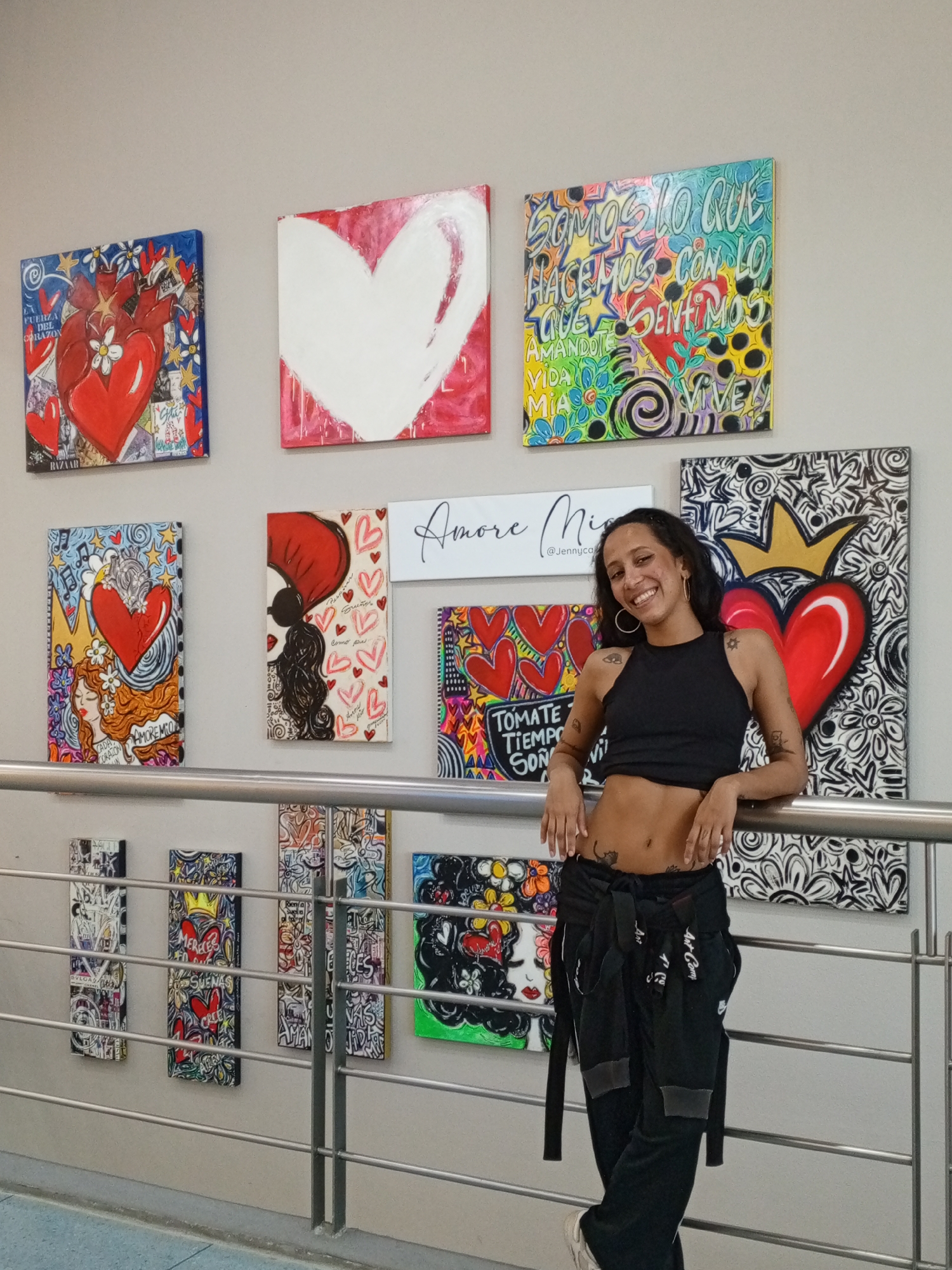 A waist-up shot of a young, fair-skinned woman with dark hair and multiple tattoos, wearing a black crop top and loose black pants, leaning against a metal railing in what appears to be an art gallery or studio. The background wall is adorned with a variety of colorful and abstract paintings, many featuring hearts. Some of the paintings feature lettering, including phrases in Spanish such as "LA FUERZA DEL CORAZON," "SOMOS LO QUE HACEMOS CON LO QUE SENTIMOS," and "TOMATE TIEMPO SON." There is also a piece with the text "Amore Mia @Jennyca" indicating the artist's name. The overall setting suggests a contemporary art space, possibly in Porlamar, Venezuela.