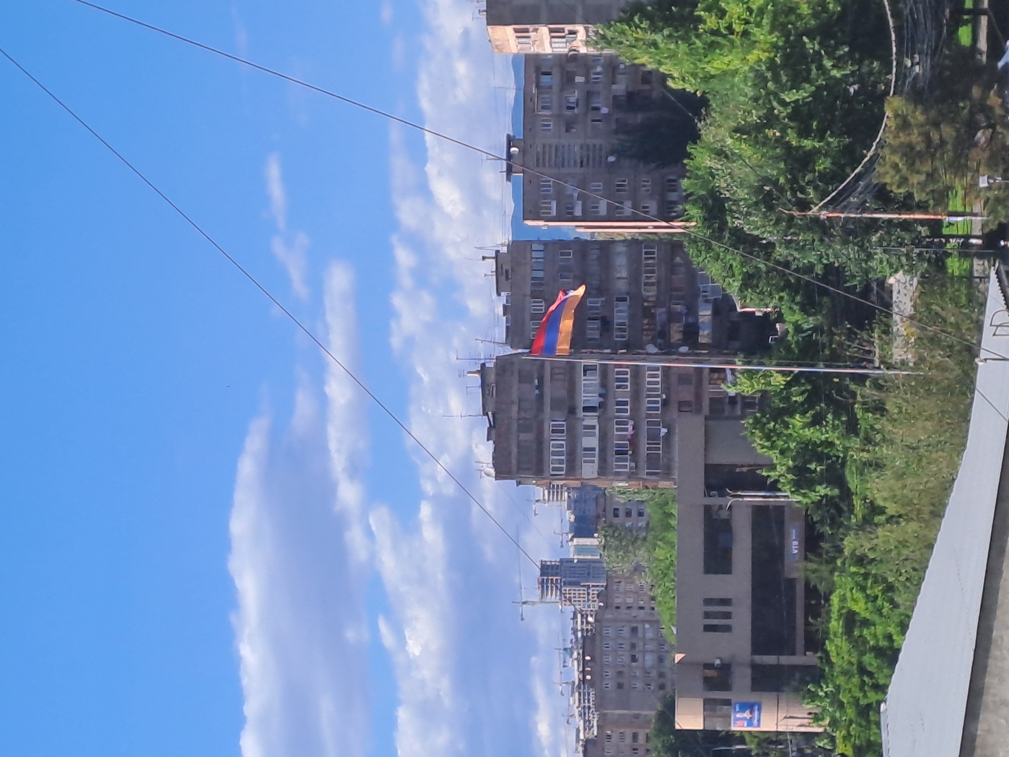 The image shows a view in Yerevan, Armenia. The Armenian flag is visible atop a building. Several multi-story residential buildings are lined up along the street. Power lines cross the sky. The sky is blue with some clouds. Green trees are present, indicating a residential or mixed-use area. A building with a "VTB" sign can be seen.