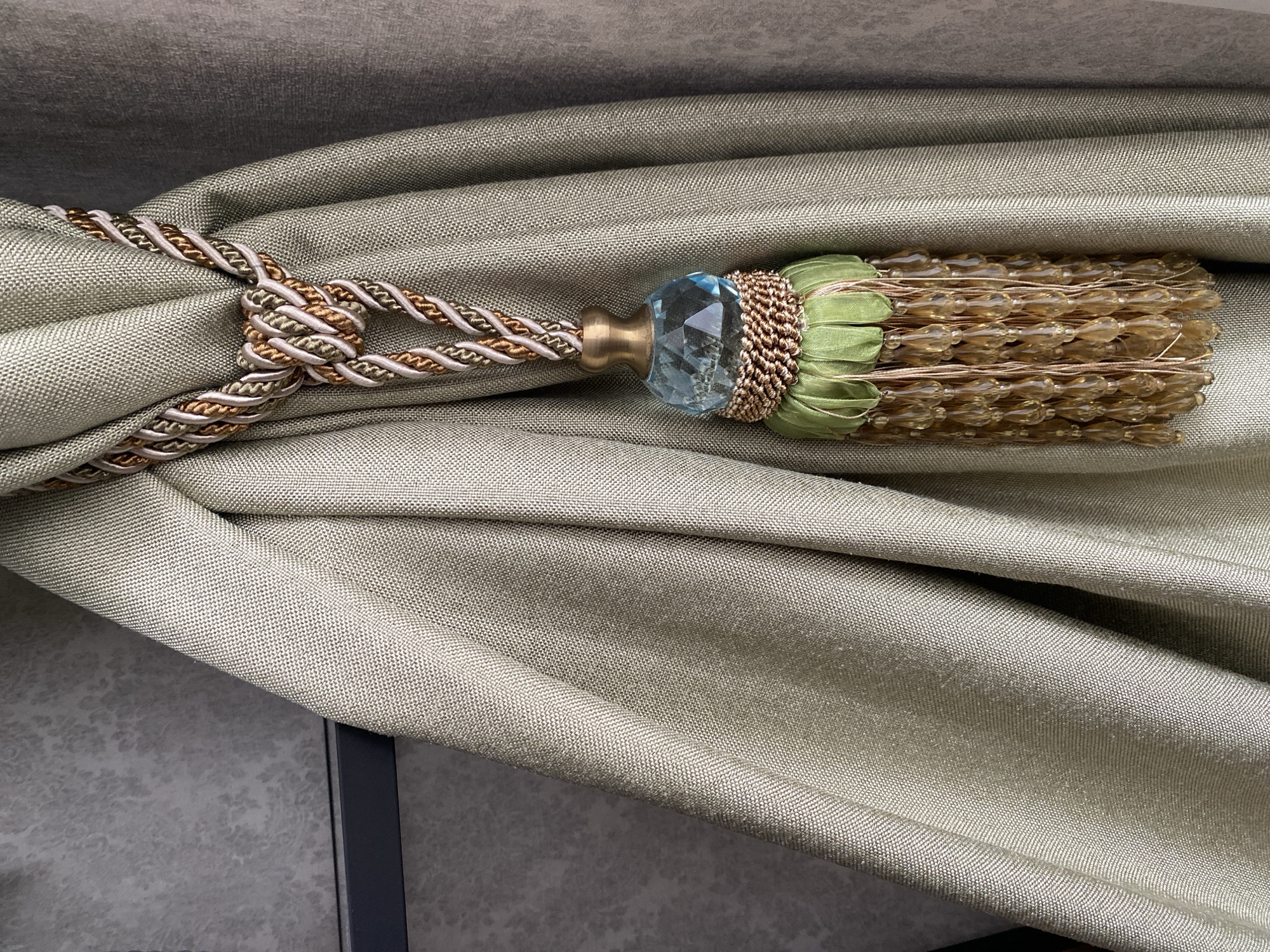 A close-up shot focuses on a decorative curtain tieback holding a draped curtain in place. The curtain appears to be made of a textured, light olive-green fabric. The tieback features a twisted cord in shades of brown, gold, and cream, looped around the curtain to gather it. The tieback tassel consists of a brass fixture, a faceted blue glass ornament, a small section of green ribbon, and multiple strands of amber-colored beads. The background includes sections of the curtain and a gray wall with a speckled pattern. There is no indication of the image's location being Izhevsk, Russia, or any other geographical information.