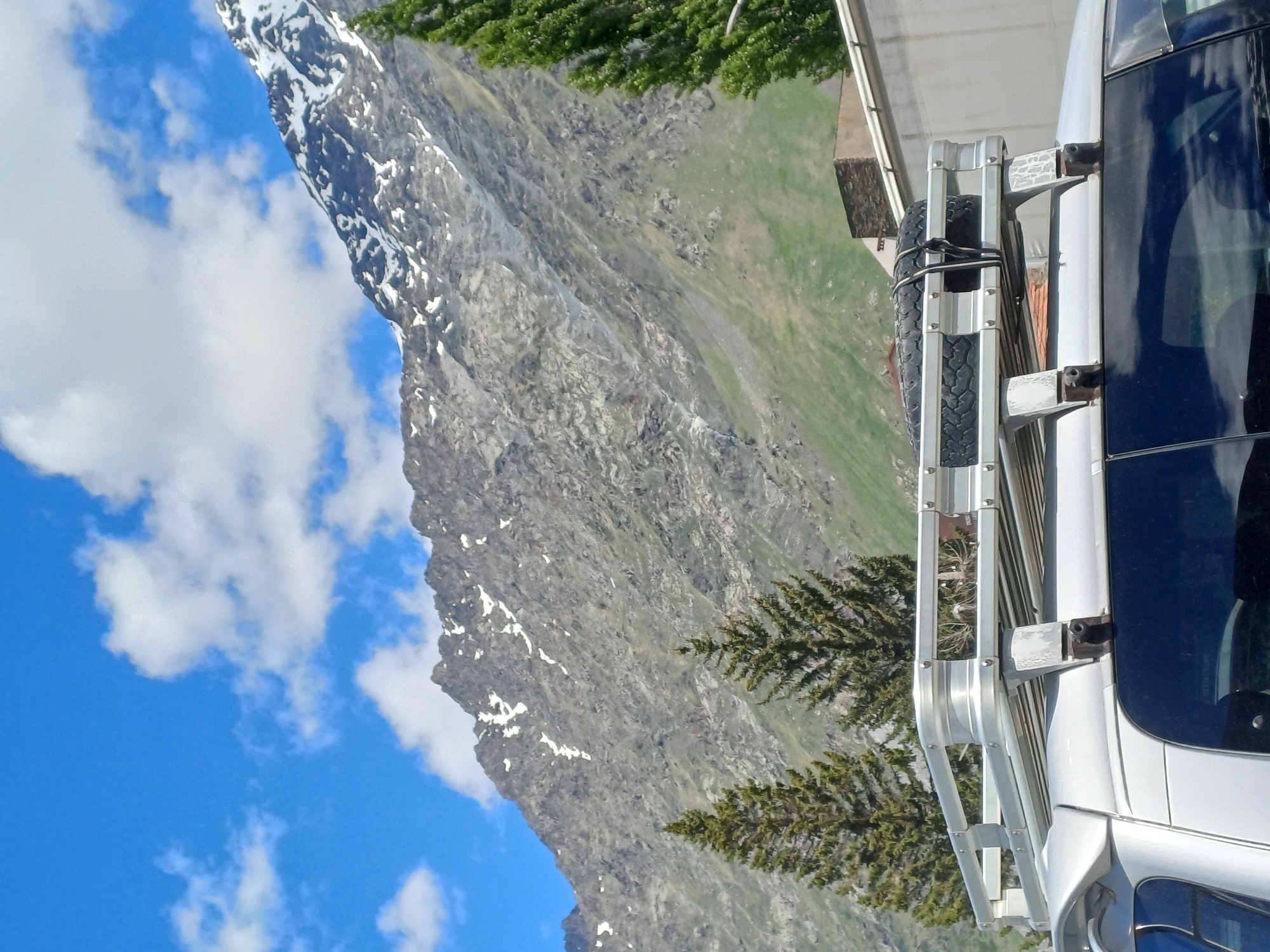 In Stepantsminda, Georgia, a silver van is parked in front of a mountain range. The van has a roof rack with a spare tire secured by black straps. Two coniferous trees are visible in the foreground, to the left and center of the image. The mountain slopes are covered in green grass and scattered patches of snow. The sky is blue with scattered white clouds. A building with a tiled roof and stone facade is partially visible behind the van.