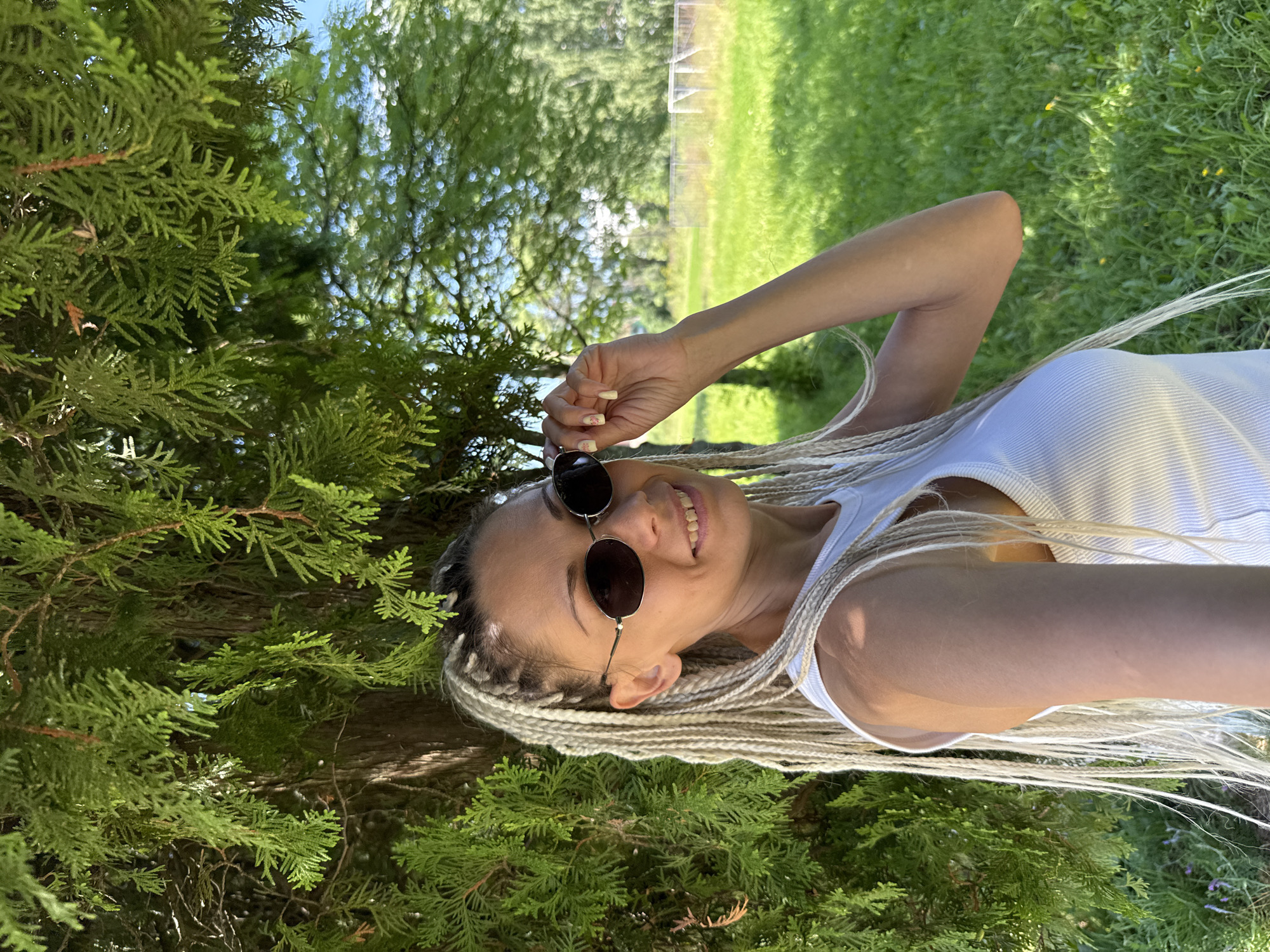 In Rusanovo, Russia, a woman with blonde braids and sunglasses is photographed outdoors. She is wearing a white tank top and is standing next to a tree. The background consists of a grassy field and trees. The woman is looking at the camera and smiling.