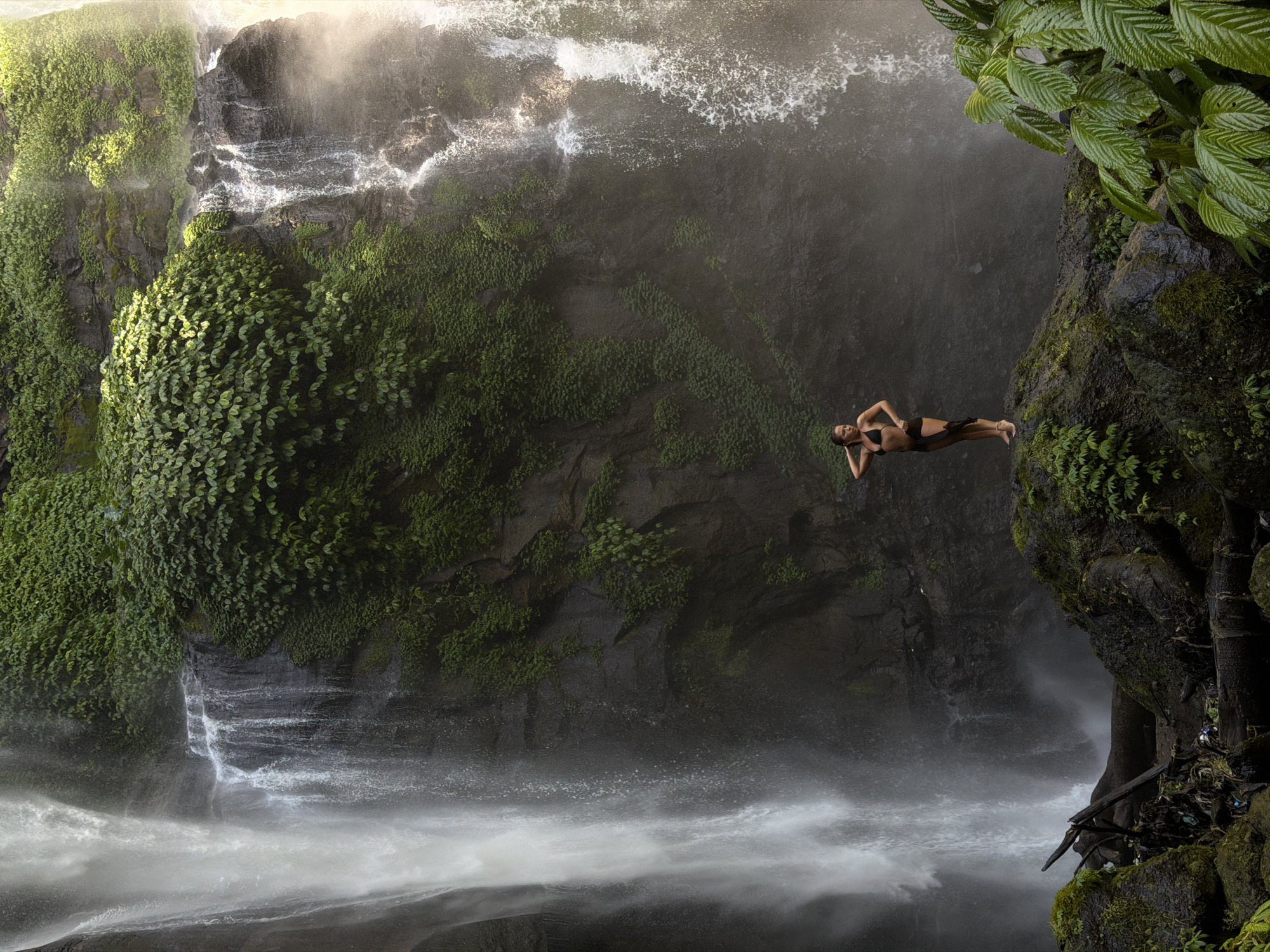 In the city of Sawan, Indonesia, a woman stands on a moss-covered rock formation in front of a large waterfall. She is wearing a black bikini and has a dark sarong tied around her waist. The waterfall cascades down a rocky cliff face that is covered in lush green vegetation. The water creates a misty spray that partially obscures the background. In the foreground, there are more rocks and some fallen branches. The scene is lit by natural light, creating a dramatic and awe-inspiring atmosphere.