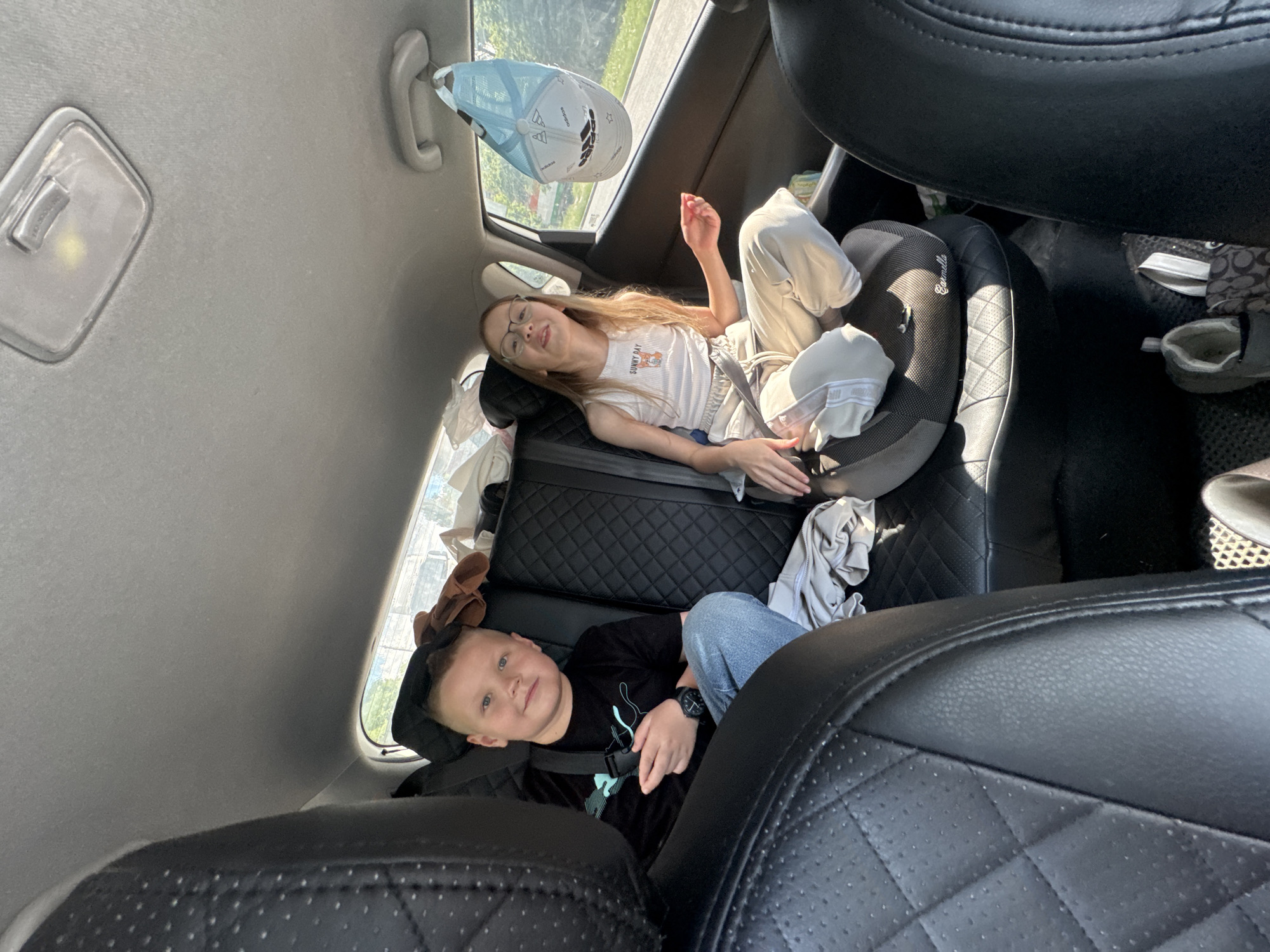 Two children are resting in the back seat of a car. A girl wearing glasses and a white tank top is reclining in a car seat. A boy in a black t-shirt is secured in a car seat below her. The car's interior features black leather seats and a light-colored headliner. A blue and white Adidas cap is hanging from the grab handle. The location could not be confirmed in Izhevsk, Russia.