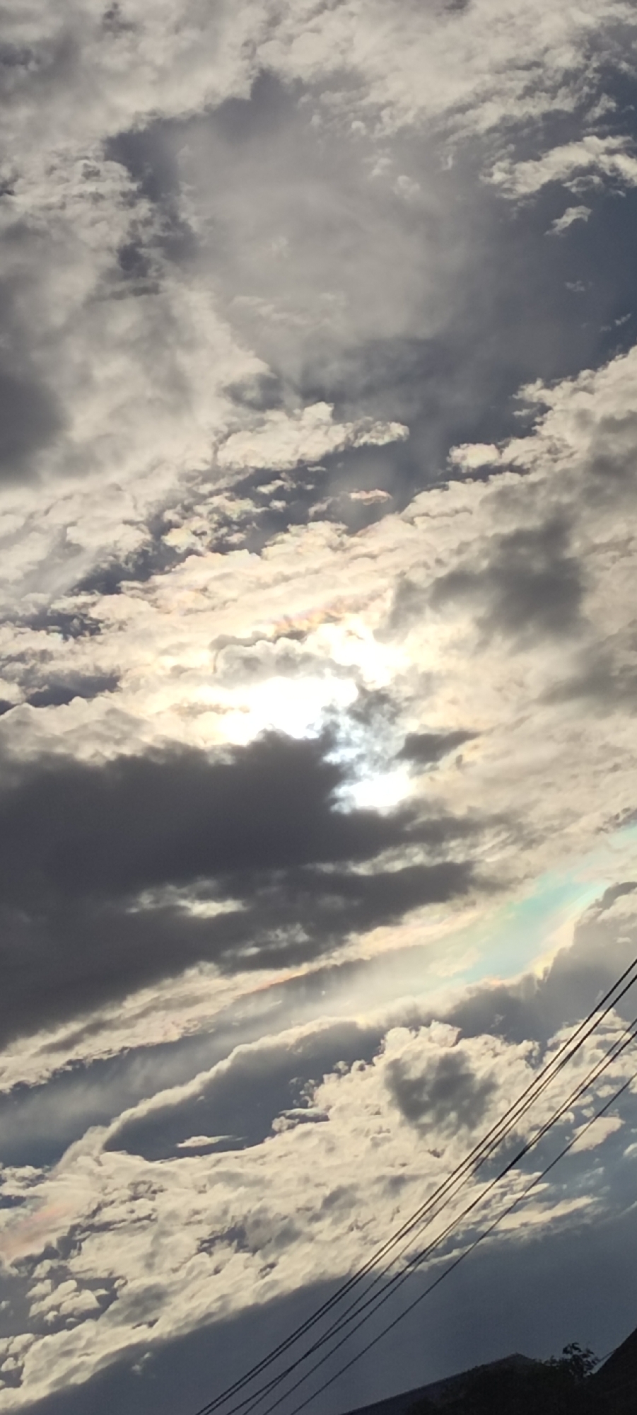 The image depicts a cloudy sky, likely captured from a ground-level perspective. The sun is partially obscured by clouds, creating a bright, diffuse light in the center of the frame. Some rainbow iridescence is visible in the clouds. In the lower right corner, power lines are visible against the sky, suggesting an urban or suburban location. The bottom edge of the frame includes vegetation and possibly a building roofline. The location is potentially Keudeteupinpunti, Indonesia, but this is not definitively verifiable from the image alone.