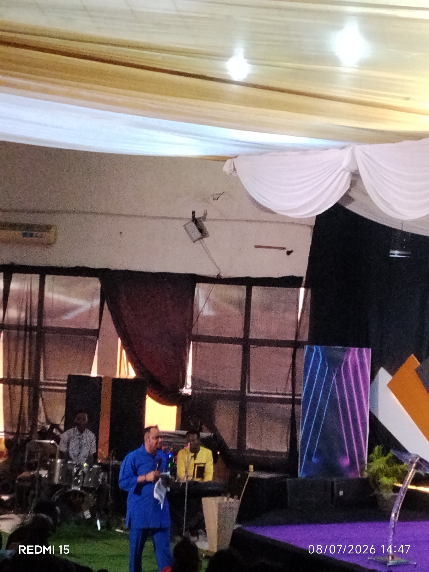 The image captures a scene at an indoor event, possibly a church service or conference, in Aba, Nigeria. A man in a vibrant blue traditional outfit is standing center stage, holding a microphone and a white cloth. He appears to be speaking or leading the congregation. To his left, a person is seated at a drum set, and to his right, another person in a yellow and black outfit is near a keyboard. Behind the main subjects, large speakers are visible, and beyond them, dark curtains hang. The stage features a decorative, illuminated structure with diagonal blue and pink lines. The ceiling is draped with light-colored fabric, and a projector or light fixture is mounted on the wall. The overall setting suggests a formal gathering with a stage setup for a performance or presentation. The text "REDMI 15" and a date and time stamp "08/07/2026 14:47" are overlaid on the image, likely indicating the device and time the photo was taken.