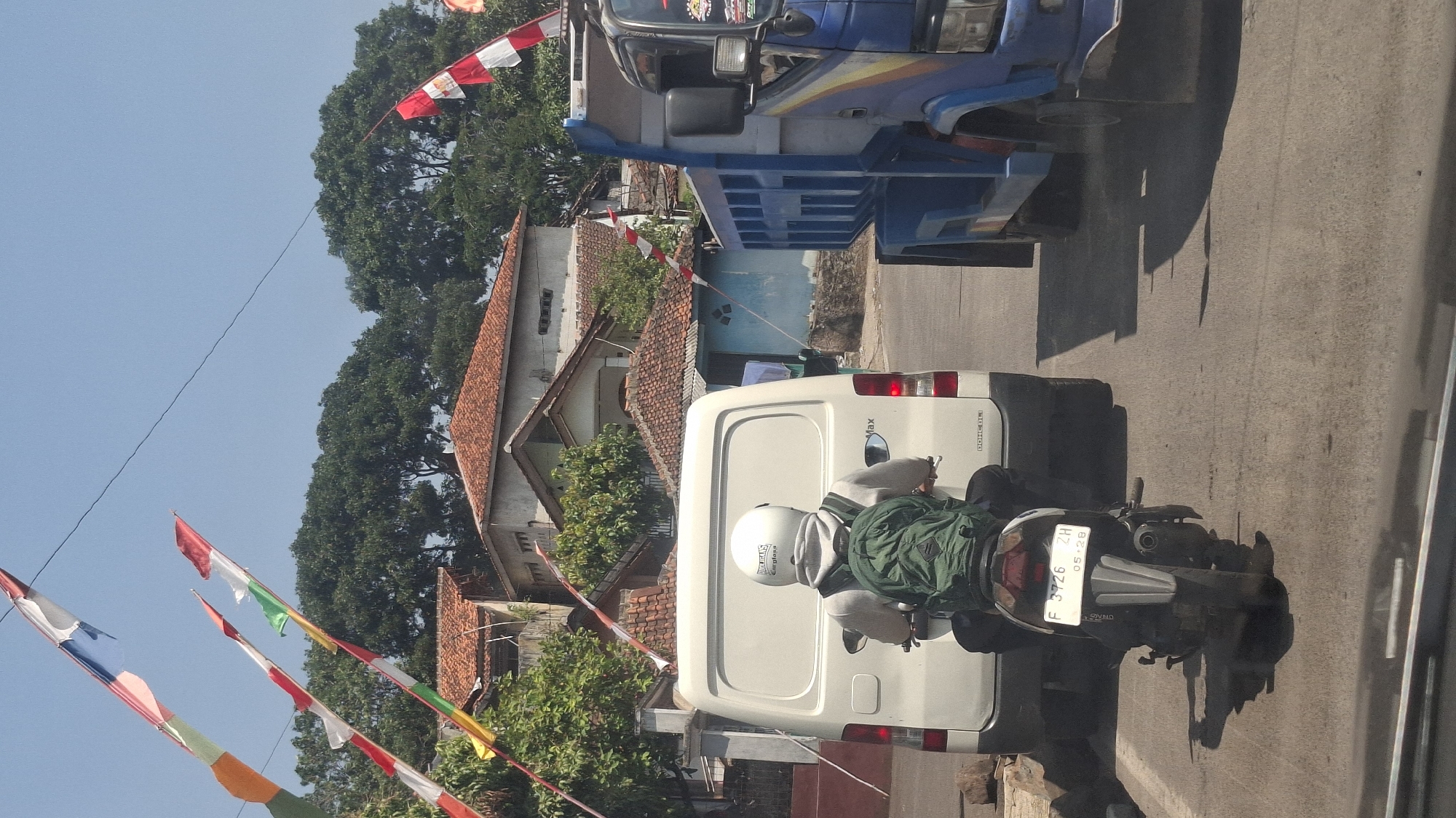 The image shows a street scene in Cilaku, Indonesia. In the foreground, a person wearing a white helmet and a green backpack is riding a motorcycle. The motorcycle has a license plate that reads "F 3726 ZH 05-28". To the left of the motorcycle is a white van, and to the right is a blue dump truck. In the background, there are several houses with red tile roofs, and lush green trees. Several flags are strung across the sky. The sky is clear and blue, indicating a sunny day. The road appears to be paved and there are some signs of wear and tear. The overall impression is of a typical street scene in a residential area of Indonesia.