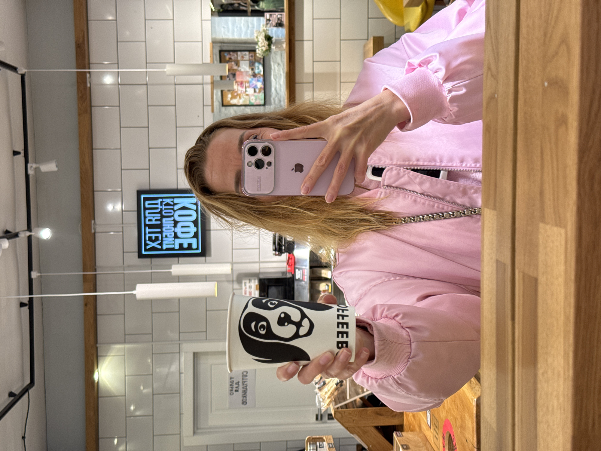 A fair-skinned individual with blonde hair, dressed in a pink satin jacket, is taking a photo of themselves in a mirror, holding a coffee cup with a black and white dog illustration on it. The individual is using a lilac-colored iPhone to capture the image. The background features a tiled wall, pendant lights, a framed picture on the wall, and a neon sign that reads "KOFFE ROLL" in Cyrillic script, indicating the location is in Russia. The sign, combined with the presence of Cyrillic text on the coffee cup, suggests the location is likely in the city of Saint Petersburg, Russia. The scene depicts a casual selfie-taking moment inside what appears to be a café.