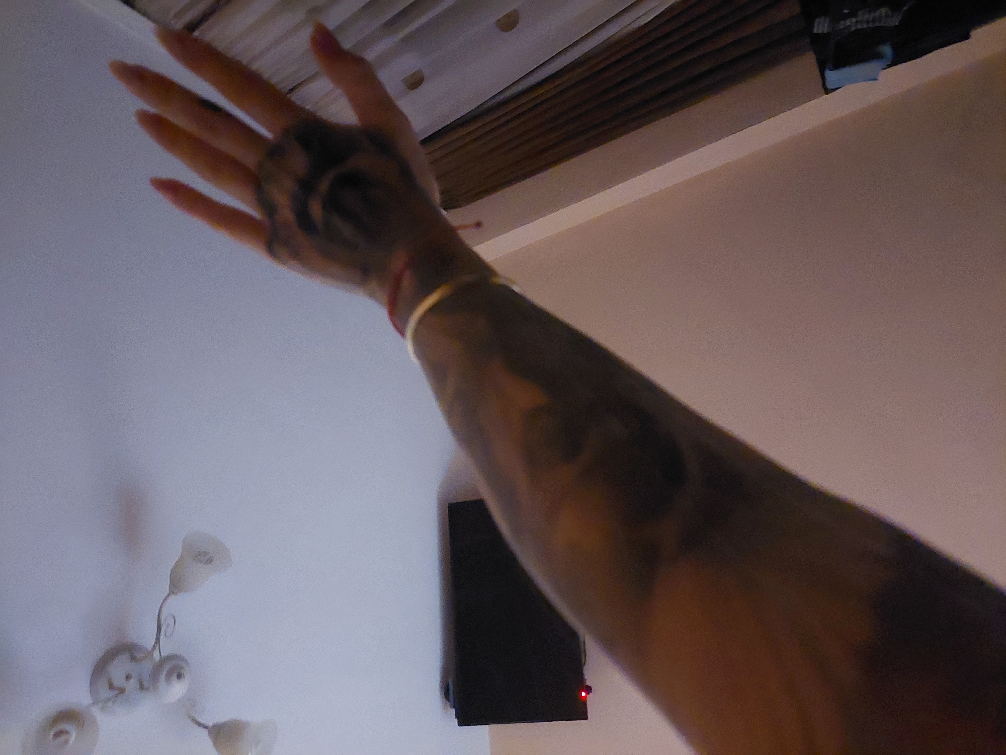 The image depicts an arm with tattoos extended upwards in an interior space. The hand, with tattoos, is outstretched. The arm is adorned with a gold bracelet and a red string.
In the background, a ceiling lamp is visible, along with what appears to be a small television mounted on a wall. The general indoor environment suggests a domestic setting. There is insufficient information to specifically determine the location as Plovdiv, Bulgaria.