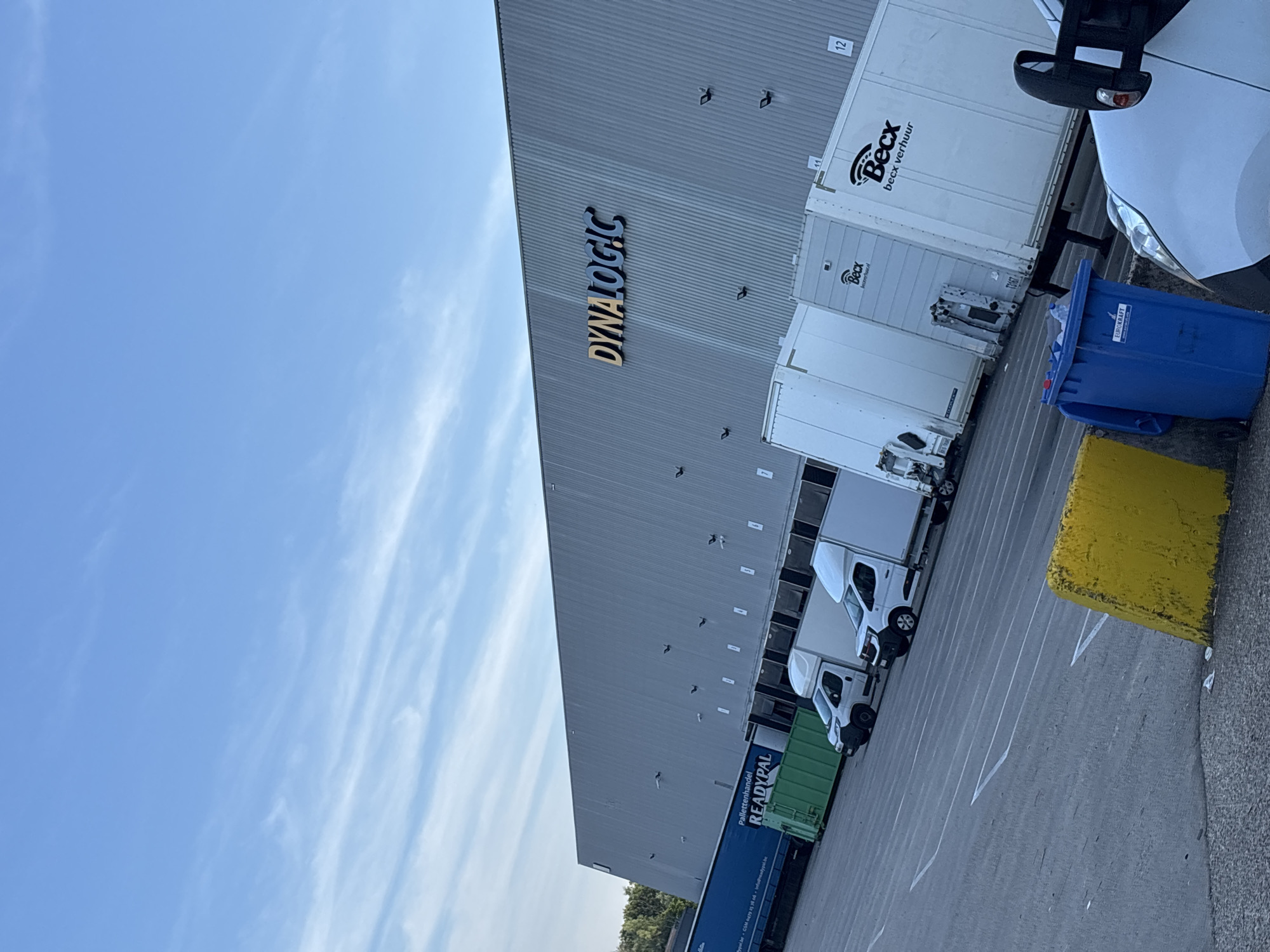 The image depicts a wide shot of a loading dock area with a large industrial building in the background. The building features the prominent signage "DYNALOGIC" in large, golden letters with a blue outline. The facade of the building is made of corrugated metal and has several loading bays.
In front of the building, several white trucks and trailers are parked, some with their doors open, suggesting active logistics operations. One trailer has "Becx verhuur" branding. A blue shipping container with "READYPAL" branding is also visible. The ground is paved asphalt, marked with white lines indicating parking spaces.
In the foreground, a bright yellow concrete barrier is present, along with a blue recycling bin labeled "EUROKRAFT." A portion of a white vehicle, likely another truck or van, is visible in the lower right corner. The sky above is a clear blue with scattered, wispy white clouds, suggesting daylight conditions.
The provided context places this scene in the city of Boom, Belgium. This indicates a commercial or industrial zone within the city, likely a distribution or logistics hub. There are no people clearly visible in the image, but the presence of vehicles and the loading bay infrastructure implies human activity related to transportation and warehousing.