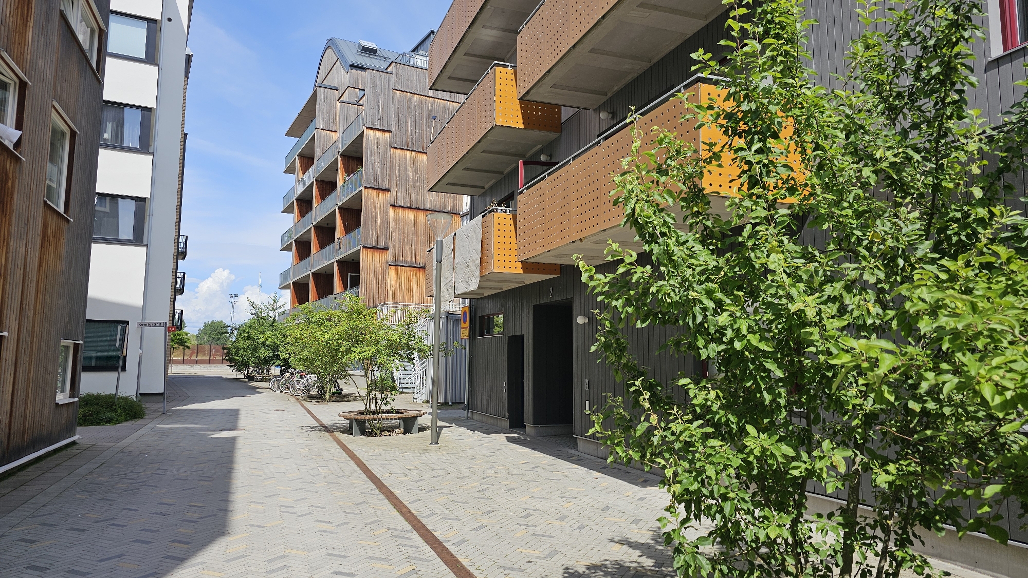 This image depicts the area outside the apartment buildings at Kärnaängen 1-15 in Linköping, Sweden, on a sunny day with a bright blue sky and scattered white clouds. The scene showcases modern residential architecture characterized by wooden facades and balconies with orange perforated detailing.
In the foreground, a tree with lush green foliage dominates the right side of the frame, partially obscuring the building behind it. The ground is paved with grey and beige interlocking bricks arranged in a herringbone pattern, with a thin red-brown line running diagonally across the walkway.
To the left, two apartment buildings are visible. The building on the far left has a wooden facade and several windows. Adjacent to it is a white building with dark-framed windows and balconies. A street sign reading "Kemigränd" is affixed to a pole between the two buildings.
In the middle ground, a building with a curved roof and multiple stories features wooden cladding and balconies. Several trees are planted along the walkway, and bicycles are parked nearby. A dark grey building with the number "2" above its entrance is situated in front of the larger wooden apartment complex. A bench with a wooden seat and a metal frame is placed near the trees.
The background shows more trees and a glimpse of what appears to be a street or open area with distant structures. The overall impression is one of a well-maintained, modern urban residential environment.