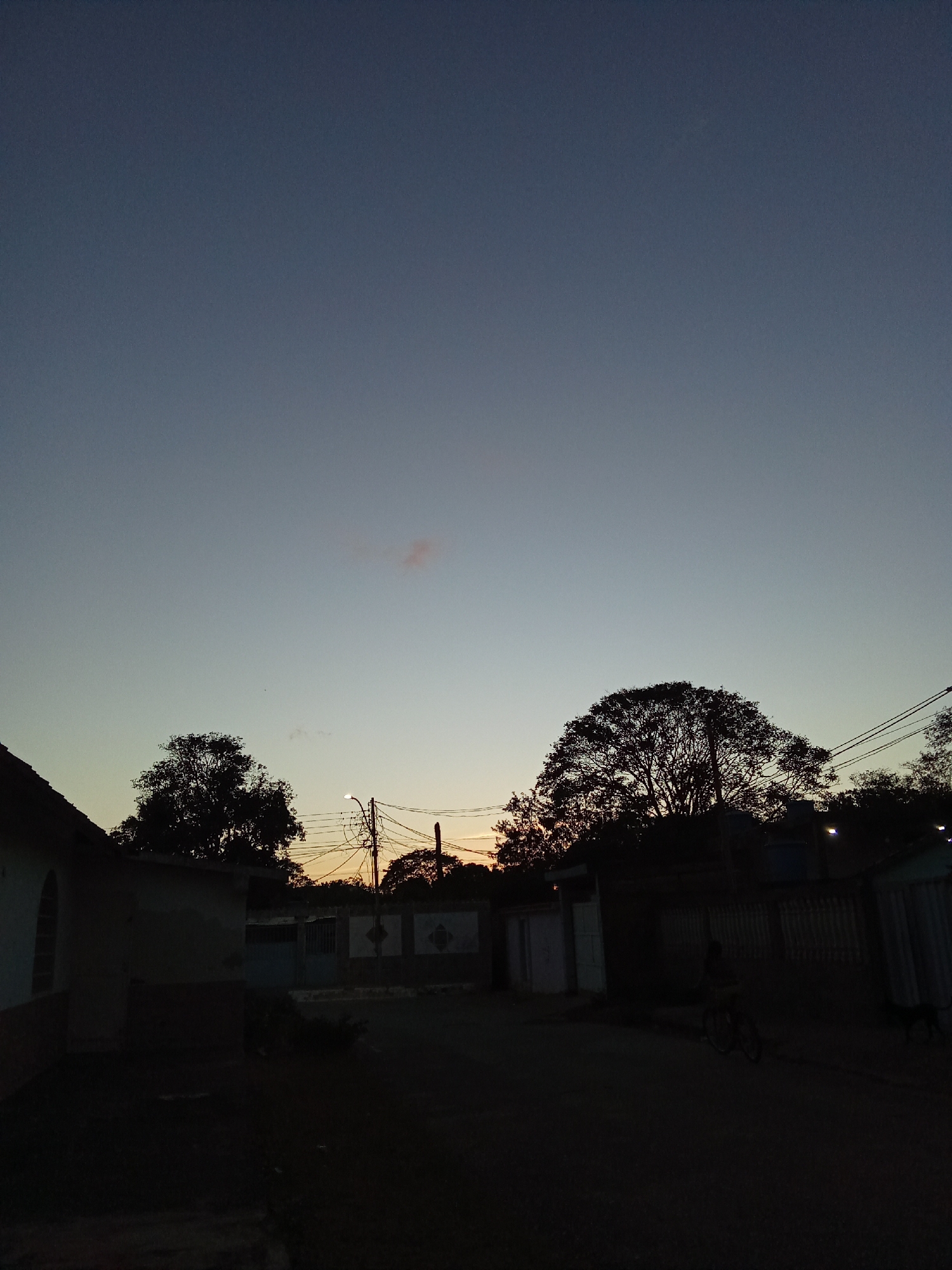 A street scene at twilight in Juan Griego, Venezuela. The sky transitions from blue to orange near the horizon, with silhouetted trees and buildings lining the road. Overhead, power lines are visible. In the foreground, the street is dark, obscuring details, though a figure on a bicycle and a dog are faintly discernible near the right edge of the frame. The buildings appear to be a mix of houses and walls, with minimal visible detail due to the low light. The overall atmosphere is calm and still, typical of a residential area at dusk.