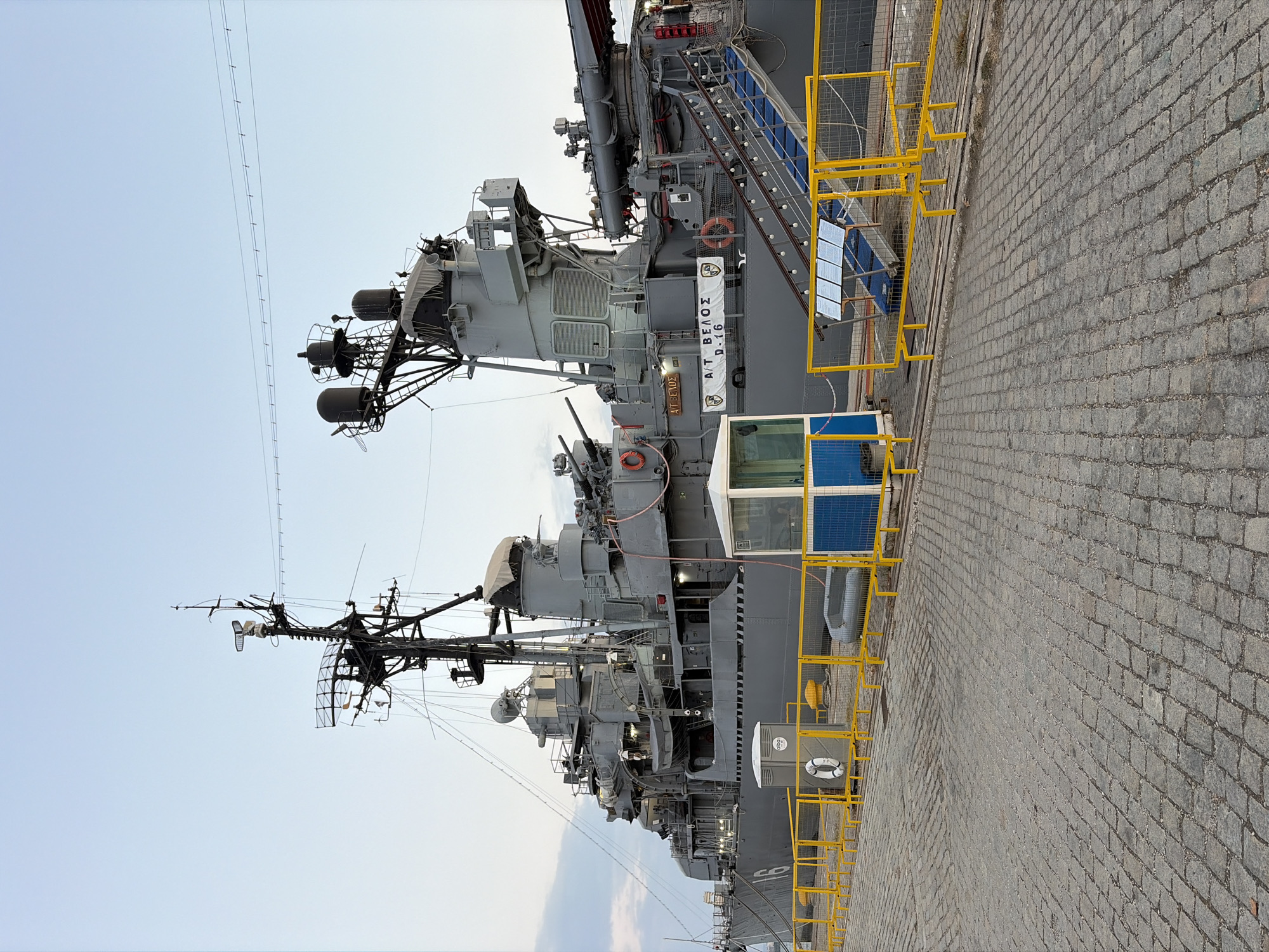 The image captures the moored warship A/T VELOS, identified by the insignia "A/T VELOS D-16" visible on its superstructure, docked at the port of Thessaloniki, Greece. The ship, painted in a matte gray, dominates the scene, with its various masts, radar arrays, and gun turrets clearly discernible. A yellow safety fence runs along the dock, delineating the boundary between the paved waterfront and the ship. A small, glass-enclosed booth with blue accents is positioned near the fence, likely serving as a checkpoint or information booth. Access to the ship is provided by a blue-carpeted gangway with illuminated railings. The cobblestone pavement in the foreground suggests a historic waterfront area. The sky above is a clear, pale blue, indicating daylight conditions. No people are visible in the frame.