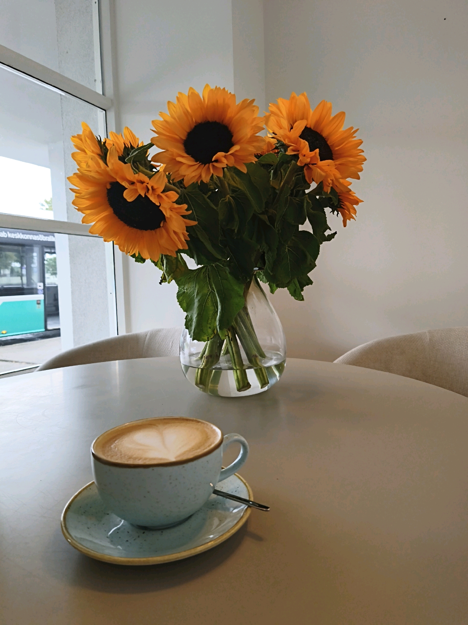 A still life photograph shows a cup of coffee with latte art, a vase of sunflowers, and a table in what appears to be a cafe. The coffee is in a light blue speckled mug with a matching saucer. The latte art forms a heart shape. The sunflowers are in a clear glass vase, with vibrant orange petals and dark centers. The vase sits on a light gray, round table. In the background, through a large window, a light-green bus is visible, suggesting an urban setting. The overall ambiance is calm and bright, with soft lighting. The location of the cafe is identified as Tallinn, Estonia.