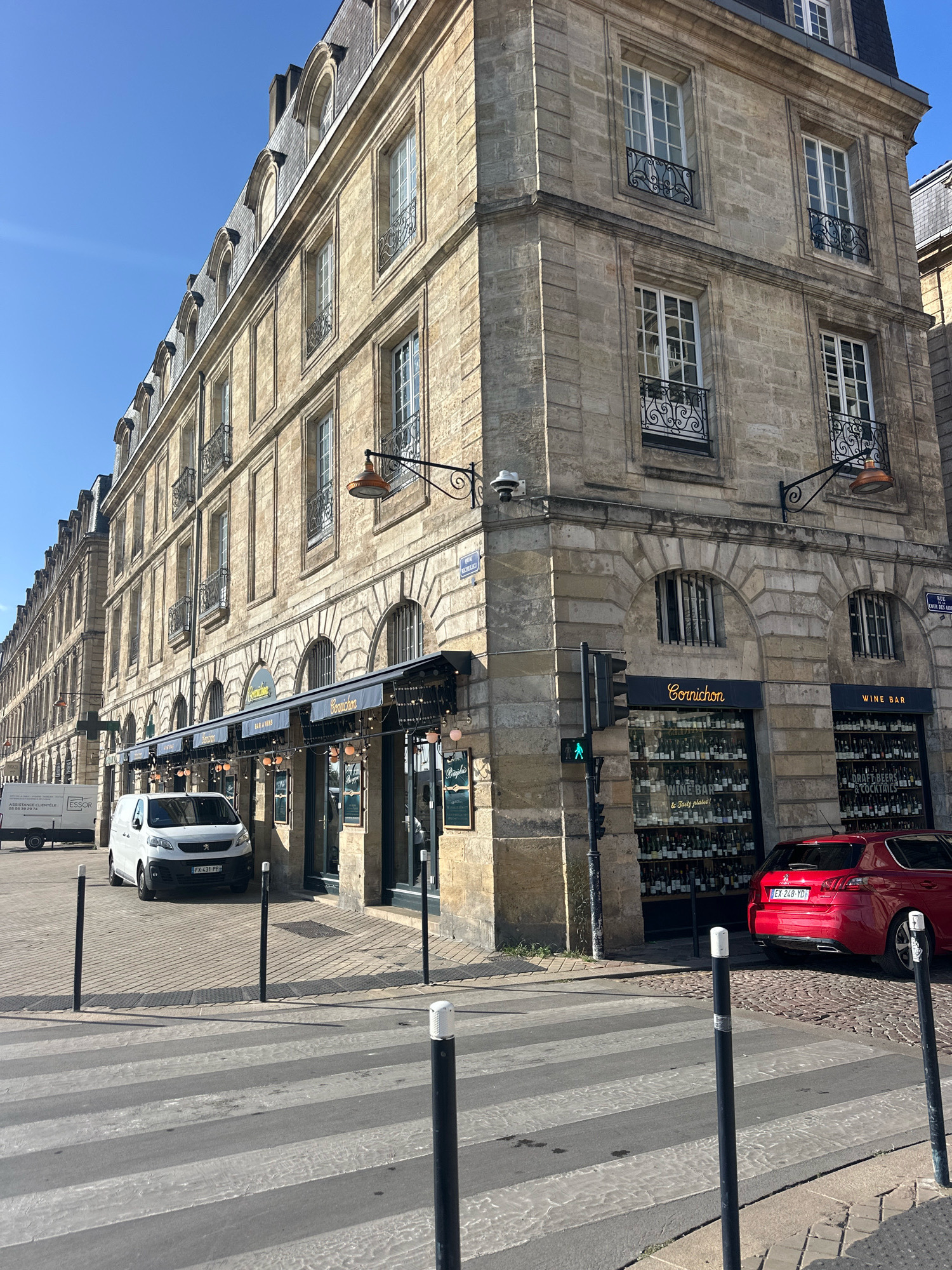 The image shows a sunlit street scene in Bordeaux, France, featuring a row of historic stone buildings. In the foreground, a pedestrian crossing is visible with black and white bollards. A white van is parked on the right side of the street, and a red hatchback car is parked in front of a wine bar. The wine bar, named "Cornichon," has large display windows filled with bottles of wine. A traffic light with a green pedestrian signal is also present. The architecture suggests a typical Parisian-style building with arched windows and ornate balconies. The sky is clear and blue.