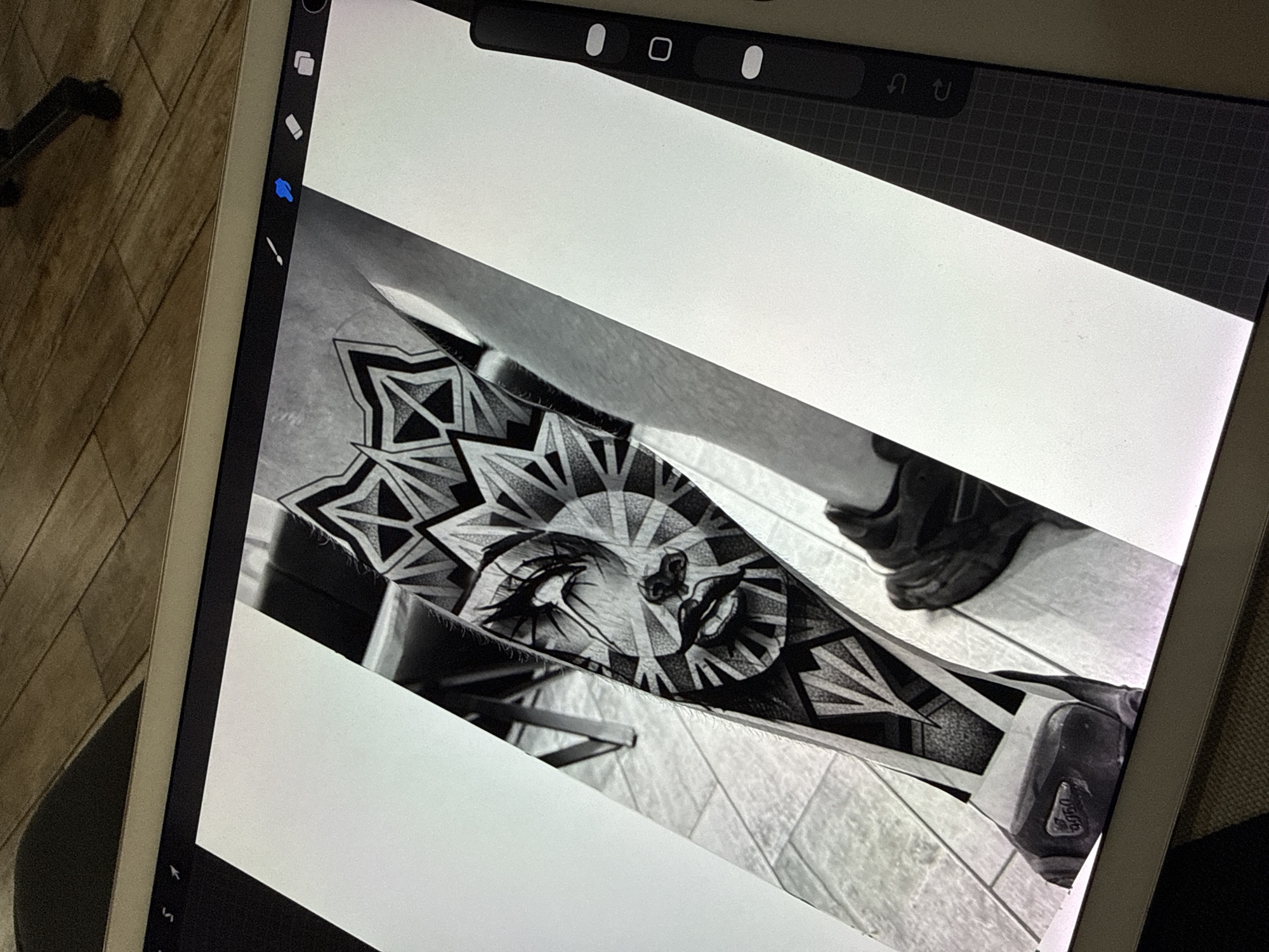 An angled shot captures an iPad or similar tablet device displaying a black and white digital artwork. The artwork depicts a highly detailed tattoo design on a person's lower leg, extending from below the knee to the foot. The central motif of the tattoo is a stylized human face with closed eyes, integrated into an elaborate geometric pattern that radiates outwards like a sunburst or mandala. This design features intricate dot work for shading and texture.
Within the artwork, parts of a person's leg and a dark-colored shoe are visible. The word "USOG" is partially legible on the sole of the shoe.
The tablet's screen prominently displays elements of a digital drawing application's user interface. On the left side, tool icons for a brush, an eraser, and a selection hand tool (highlighted in blue) are visible. At the top right of the screen, undo and redo arrows are present.
The tablet is positioned over a light-colored, tiled floor, which also forms the background within the displayed artwork. The overall scene suggests an indoor setting, possibly a studio or home, where digital art is being created or reviewed. The image is composed from a slightly elevated and tilted perspective, focusing on the tablet and its contents.