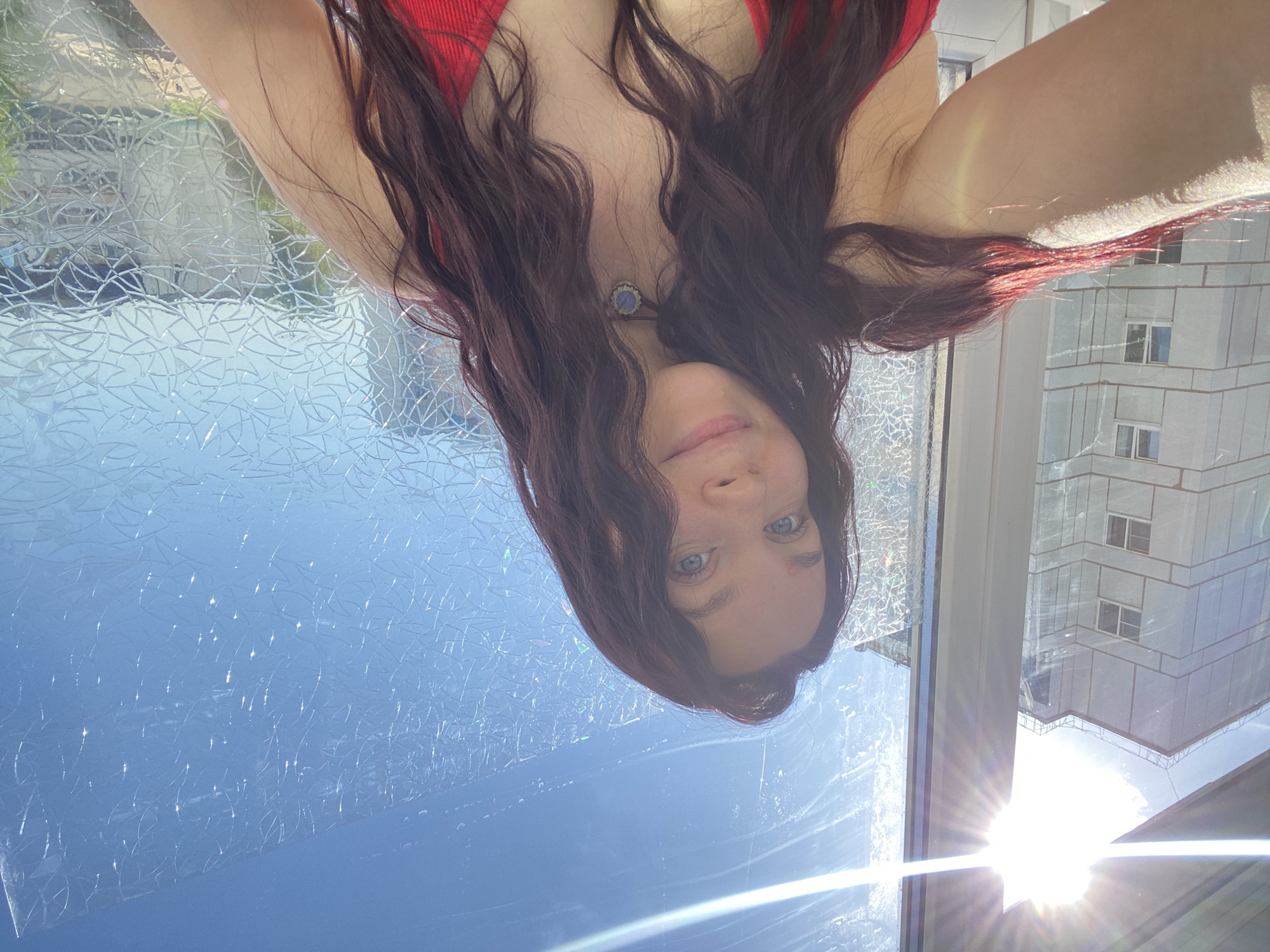 A selfie captured through a window in Chaikovsky, Russia. The window is treated with a decorative film that distorts the view outside. Reflected in the window are a building and sunlight. The selfie features a woman with long, wavy, auburn hair and blue eyes, wearing a red top and a necklace. The orientation of the image is upside down.
