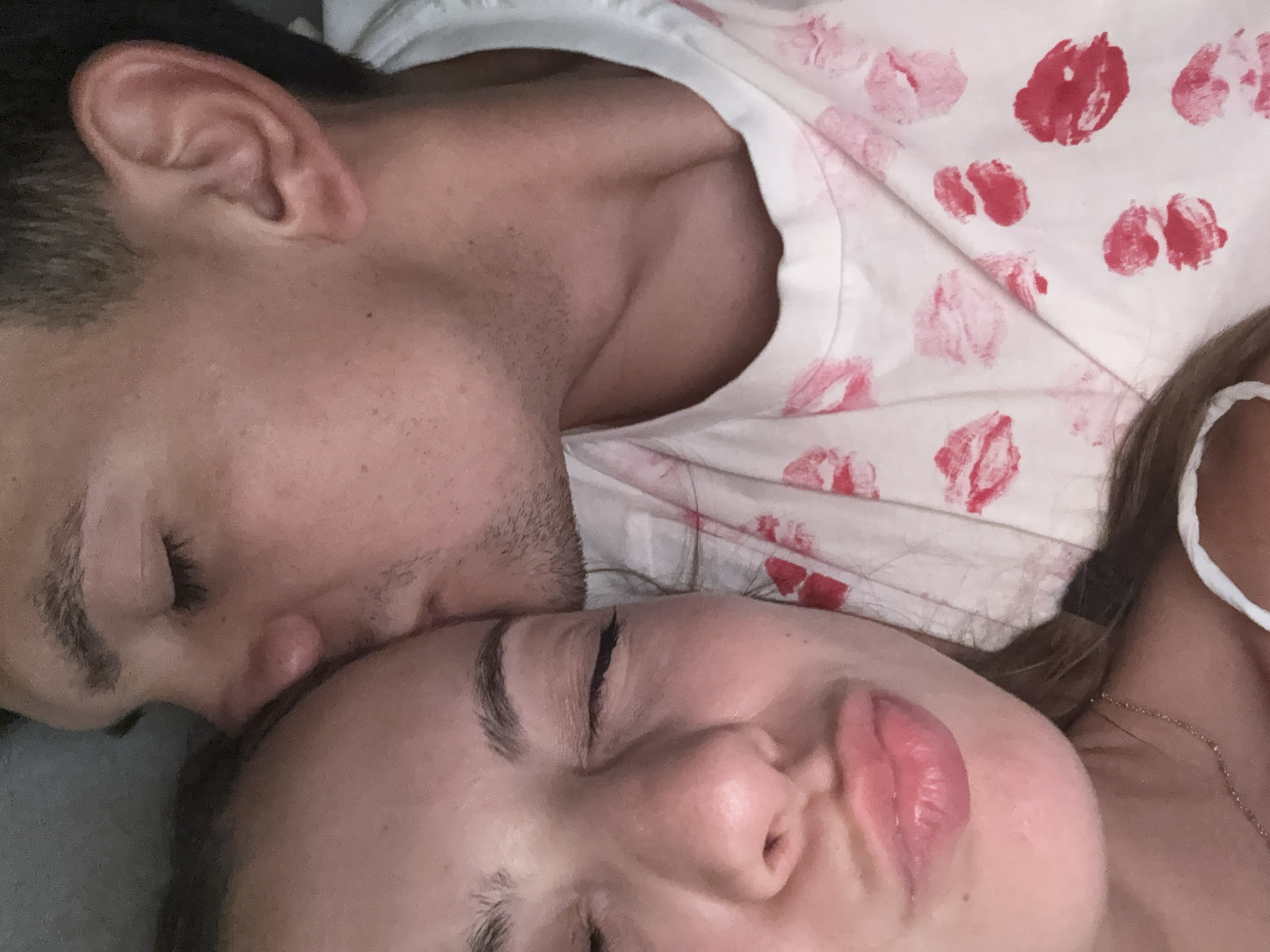 This is a close-up, indoor, eye-level photograph of a young couple embracing. The male subject is kissing the female subject on her forehead. Both individuals appear to be in their late teens or early twenties. The female subject is positioned in the foreground, facing the camera with her eyes closed and lips pursed. She has long, dark hair and is wearing a thin necklace. The male subject is slightly out of focus in the background, his face turned towards the female. He has short, dark hair and is wearing a white t-shirt with a pattern of red lipstick kisses. The background is indistinct, but suggests an indoor setting, possibly a bed or couch, given the soft lighting and close proximity of the subjects. The image does not contain any recognizable landmarks or elements that would indicate its location in Sofia, Bulgaria.