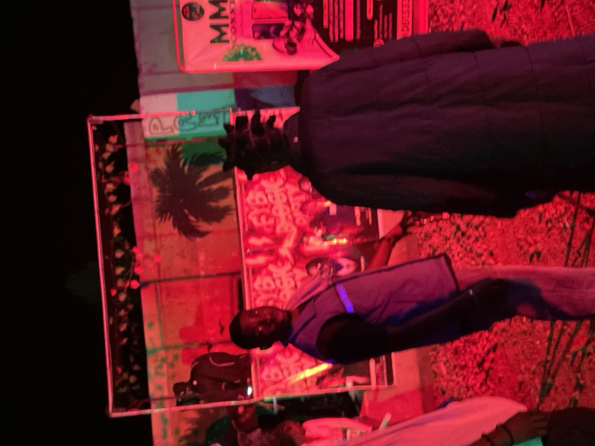 The image depicts a nighttime outdoor scene bathed in intense red lighting, possibly from stage or event lighting. In the foreground, a young Black male wearing a dark puffer jacket is seen from behind, facing away from the camera. To his left, another young Black male is facing the camera, wearing a light purple vest over a dark garment and light-colored pants. He is looking directly at the viewer with a neutral expression. Behind these individuals, a backdrop displays graphics and text, including the words "FYB BLOCK PARTY" with a tropical palm tree silhouette. To the right, a banner with the text "MM CONNECTED" is visible. The ground appears to be covered with debris, possibly gravel or leaves. The overall atmosphere suggests a lively event or party taking place in Durumi, Nigeria.