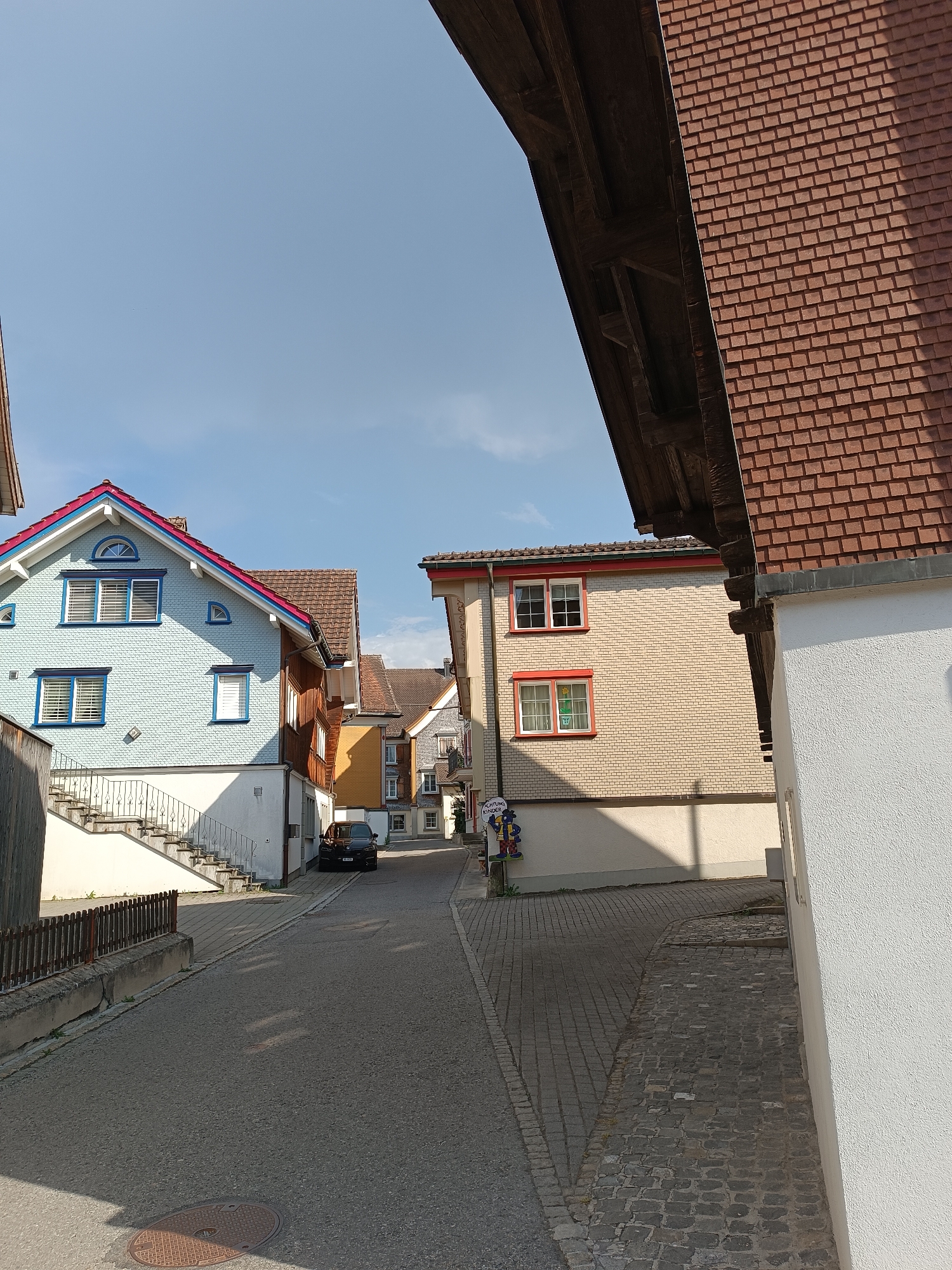 This is an eye-level, medium shot captured on a sunny day, looking down a narrow, paved street in the city of Appenzell, Switzerland. The street is lined with traditional Swiss architecture. On the left, a light blue house with a red and blue trim roof stands out. It features a patterned facade, white-framed windows with blue shutters, and a set of white stairs with a metal railing leading up to its entrance. To the right of this house is a darker wooden building. Further down the street, other buildings are visible, including a light-colored brick house with red-trimmed windows. A dark-colored car is parked on the right side of the street. The street itself is paved with asphalt, with cobblestone sections visible on the sides. The sky is clear blue, with a hint of clouds. Overhead, the wooden structure of a large building with a red tile roof juts into the frame from the top right, casting a shadow on the scene below. The overall impression is of a quaint and well-maintained village street.