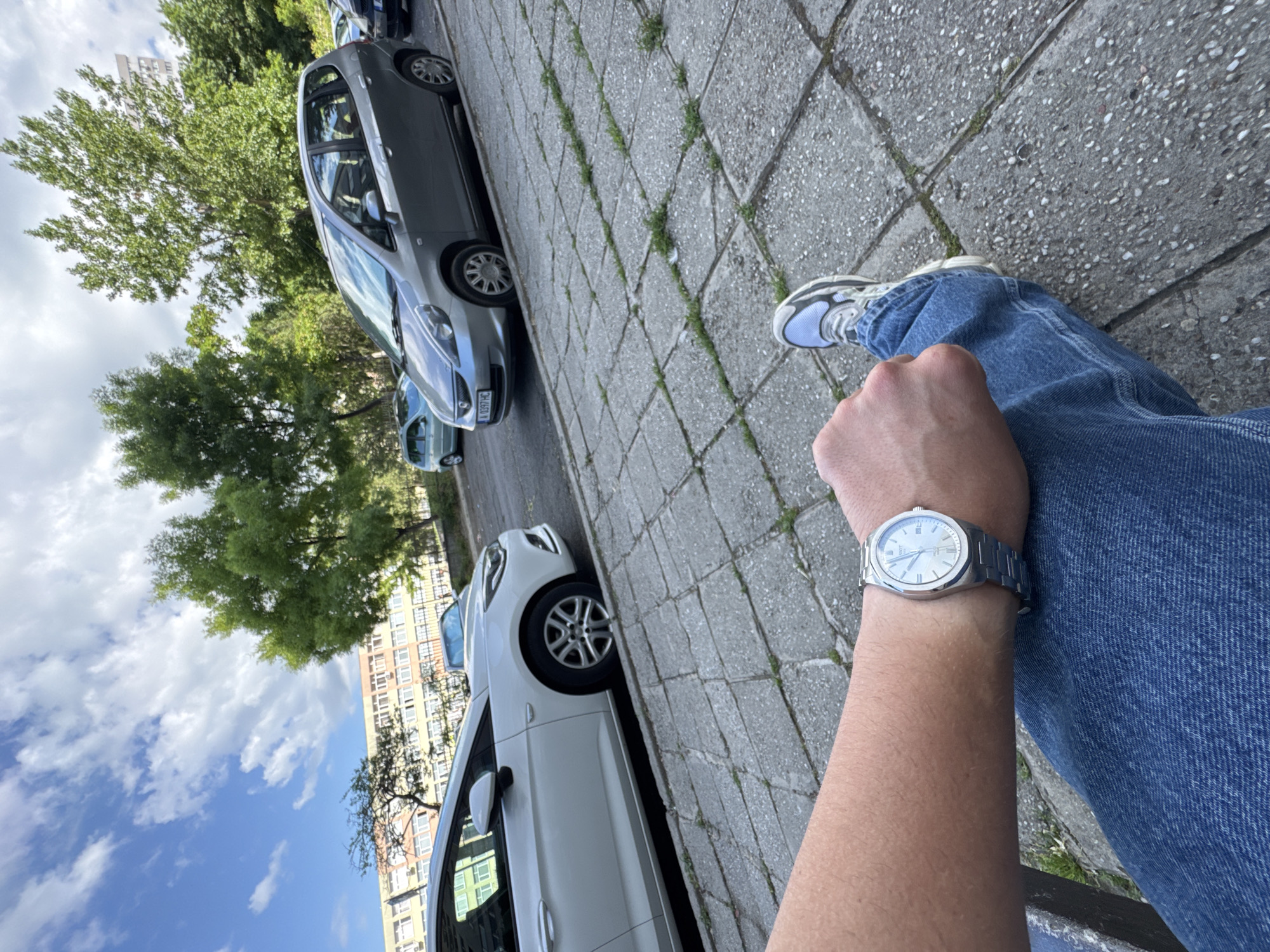 The image is taken at an angle from a first-person perspective, showing a person sitting outdoors on a sunny day. The person is wearing blue jeans and a silver wristwatch on their left wrist. Their left leg is partially visible, with a white and gray sneaker. The ground is paved with gray, square-shaped concrete tiles, with some weeds growing in the cracks. Several cars are parked on the street to the left. These cars are predominantly silver and white, with one car's license plate partially visible. Trees with green foliage are visible in the background, along with a tall building with multiple windows and a cloudy blue sky. Based on the visible license plate (ZH-6601 HC), the location is likely Burgas, Bulgaria.