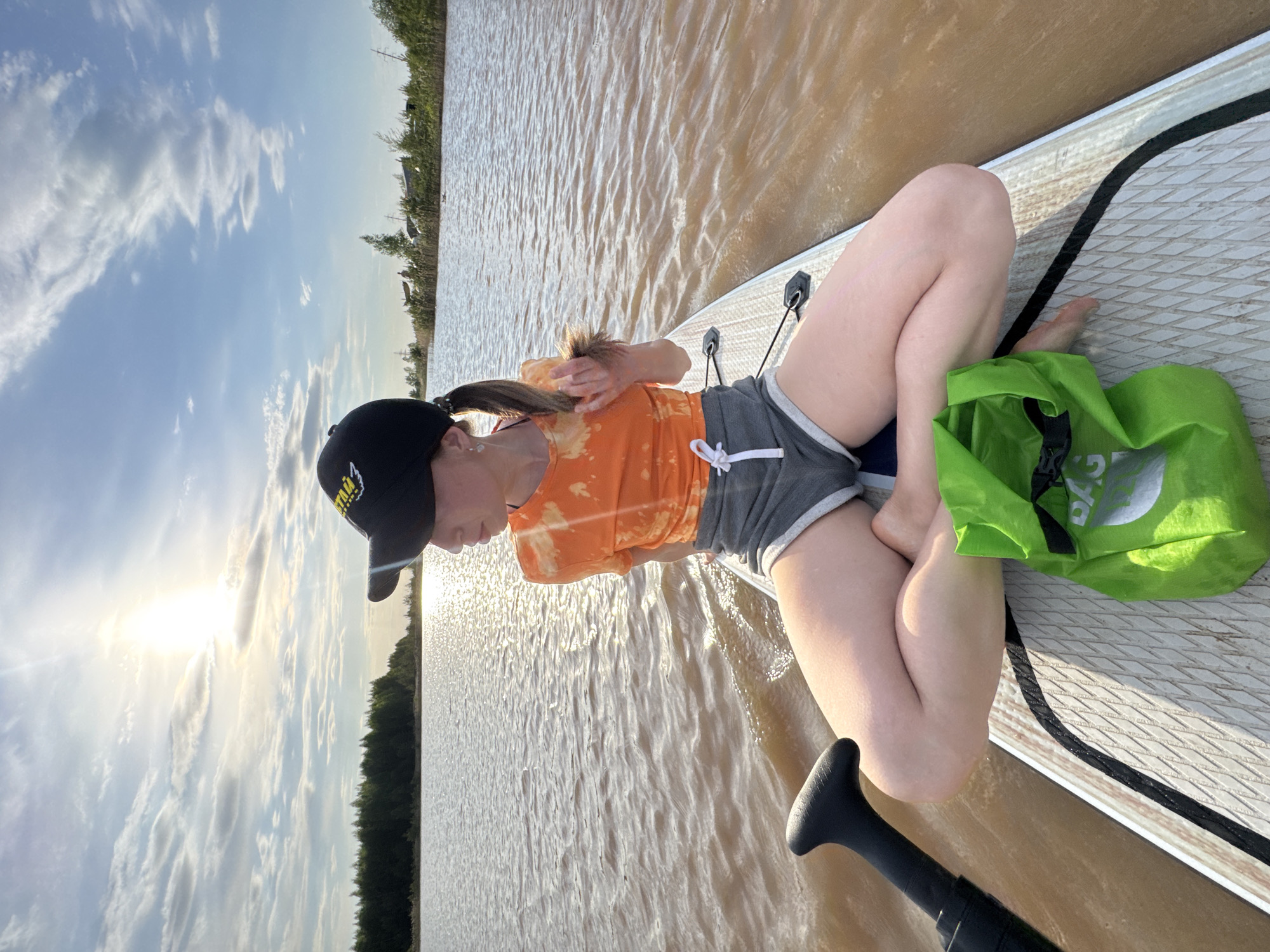 A medium, horizontally-oriented shot shows a Caucasian woman kneeling on a paddleboard in a body of muddy water under a partly cloudy sky. The woman is facing away from the camera and toward the left. She wears a black baseball cap, an orange and white tie-dye t-shirt, and gray shorts. A green bag sits to her right. A paddle is partially visible at the bottom left of the frame. The shoreline is visible in the background. The sun glares in the distance to the left, reflecting on the water. Based on the landscape and muddy water, the location appears to be in a rural area of Russia. Noviy is likely incorrect.