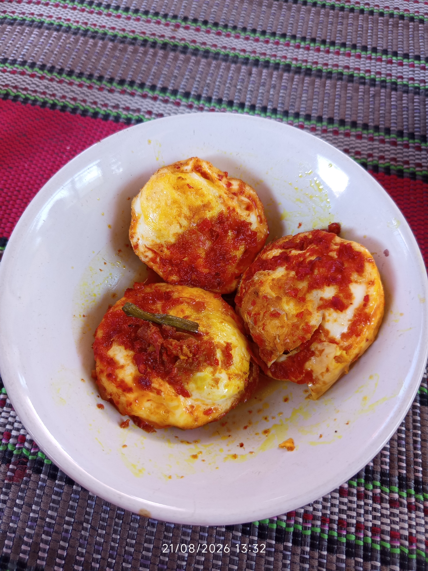 A close-up, overhead shot captures three halves of hard-boiled eggs, coated in a vibrant red chili sauce, nestled in a white ceramic bowl. The eggs have been cut in half horizontally, revealing the white and yolk interiors, with the chili mixture generously applied to the top surface. One egg half has a small, thin green garnish resting on the sauce. The bowl sits on a textured placemat, which exhibits a woven pattern with horizontal stripes in shades of red, white, black, and green. The background is a blurred continuation of the placemat. The lower right corner of the image displays a timestamp: "21/08/2026 13:32". The scene appears to be an indoor setting, possibly a restaurant or home in Canggu, Indonesia, given the typical presentation of local cuisine. There are no people visible in the frame.