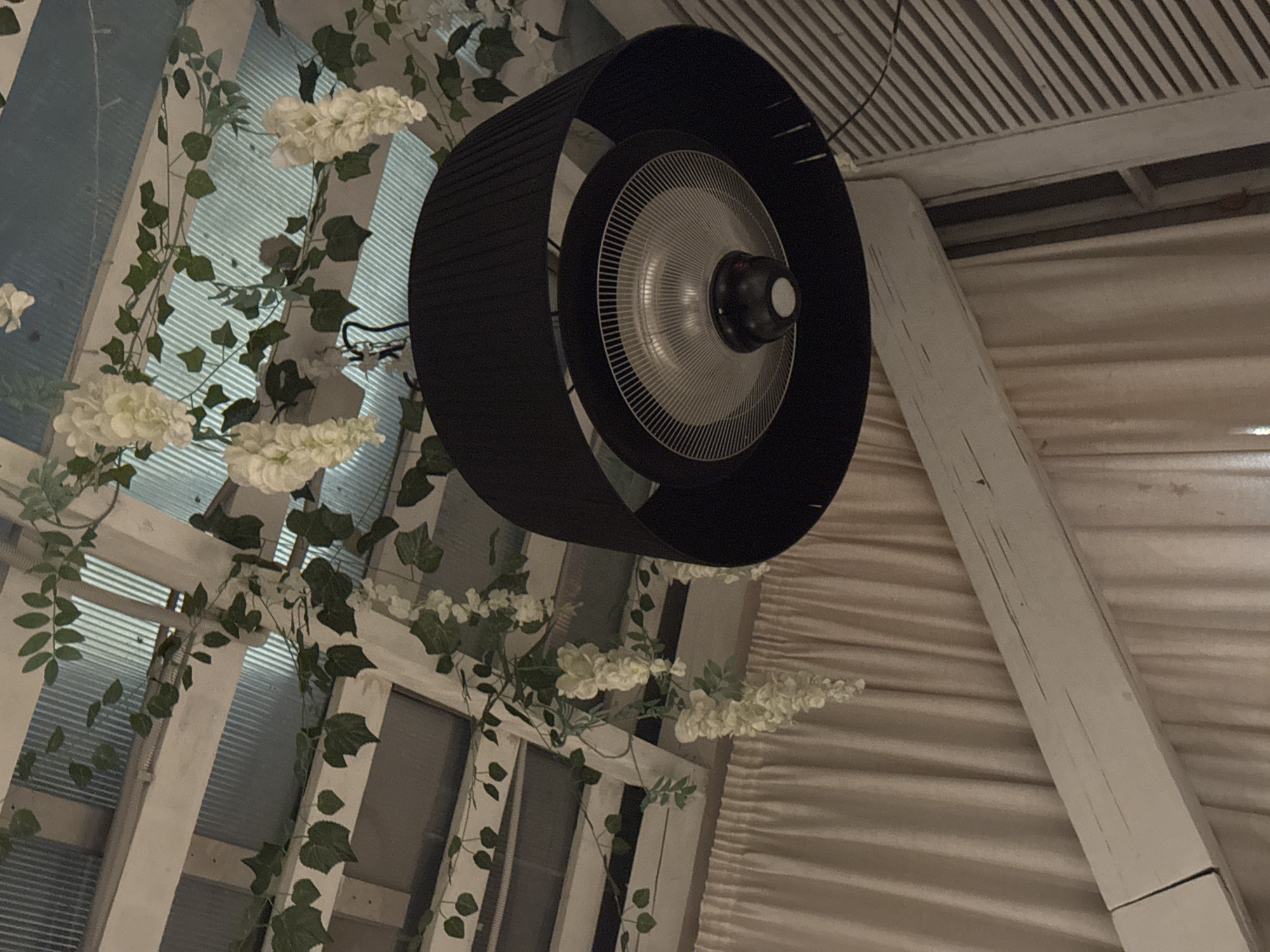 The image captures an indoor or semi-outdoor setting illuminated by artificial light, suggesting evening or nighttime. The central subject is a large, black, cylindrical ceiling-mounted heater or fan, featuring a metallic grille and a central heating/fan element. It is suspended from a white slatted ceiling structure.
To the left, artificial ivy vines with clusters of white flowers are decoratively draped over white wooden lattice work, possibly a window frame or part of a pergola. Behind the lattice, translucent ribbed panels are visible, likely forming part of the ceiling or a skylight. To the right, a prominent white wooden beam extends upwards, with light-colored, possibly fabric, drapes or wall coverings visible in the background, hinting at a tent-like structure or an enclosed patio.
No people are present, and no specific activity is taking place. The combination of the heater, decorative artificial plants, and the structural elements suggests a cozy, enclosed or semi-enclosed space, such as a covered patio, conservatory, or a restaurant/cafe seating area, designed for comfort and aesthetic appeal. The scene evokes a calm and relaxed atmosphere. There is no visible text in the image. Given the location context of Perm, Russia, the presence of a heater is practical for such an environment.