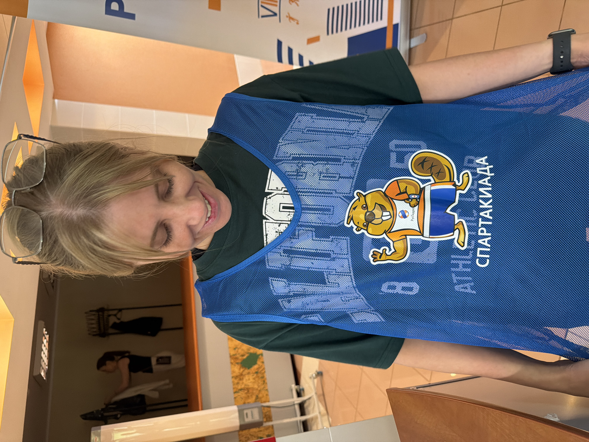 A young woman with blonde hair pulled back and wearing glasses over her eyes smiles as she looks down at a blue mesh athletic bib she is wearing. The bib has a graphic of a cartoon beaver wearing an orange shirt and blue shorts, with the words "СПАРТАКИАДА" in Cyrillic text below it. The woman is wearing a dark green t-shirt underneath the bib. In the background, two women are visible in a locker room area, with clothing racks holding jackets and coats. The environment appears to be indoors, possibly at a sports facility or school. The lighting is bright, with yellow accents on the ceiling. The woman is positioned in the foreground, with the background slightly out of focus.