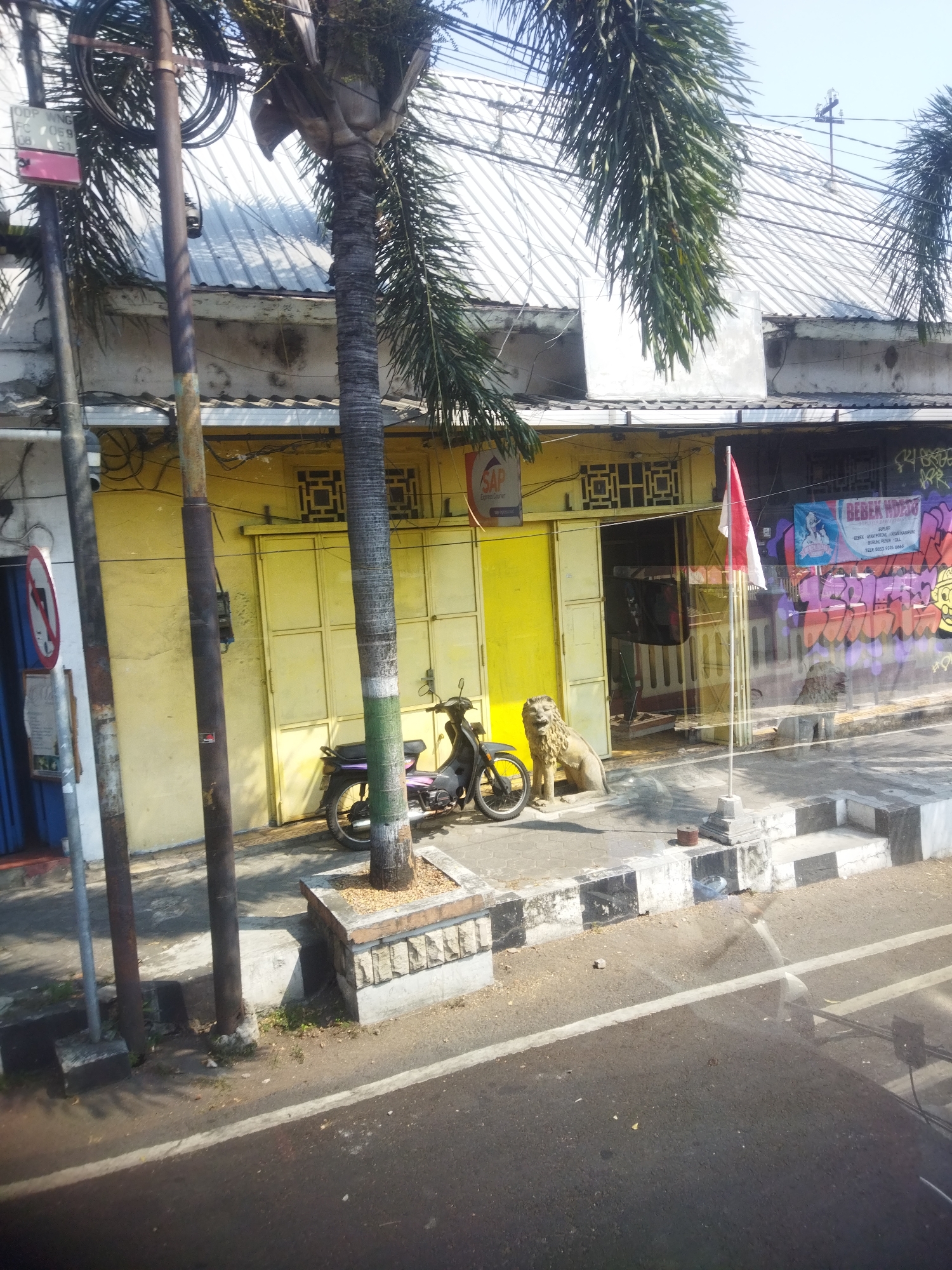 The image depicts a street scene in Giripurwo, Indonesia, featuring a yellow building with a white metal roof. Several palm trees with dark, textured trunks are visible. In front of the building, a black motorcycle is parked. To the right of the motorcycle, a concrete statue of a lion sits on the sidewalk, and further to the right, a flag pole with a red and white flag is planted. The building's facade has multiple windows with a geometric lattice design and a large, yellow sliding door. A sign with the text "SAP Express Courier" is attached to the building. The sidewalk is bordered by a curb with black and white stripes. The street in the foreground is paved and appears to be a road. Graffiti is present on the wall of an adjacent structure to the right of the yellow building.