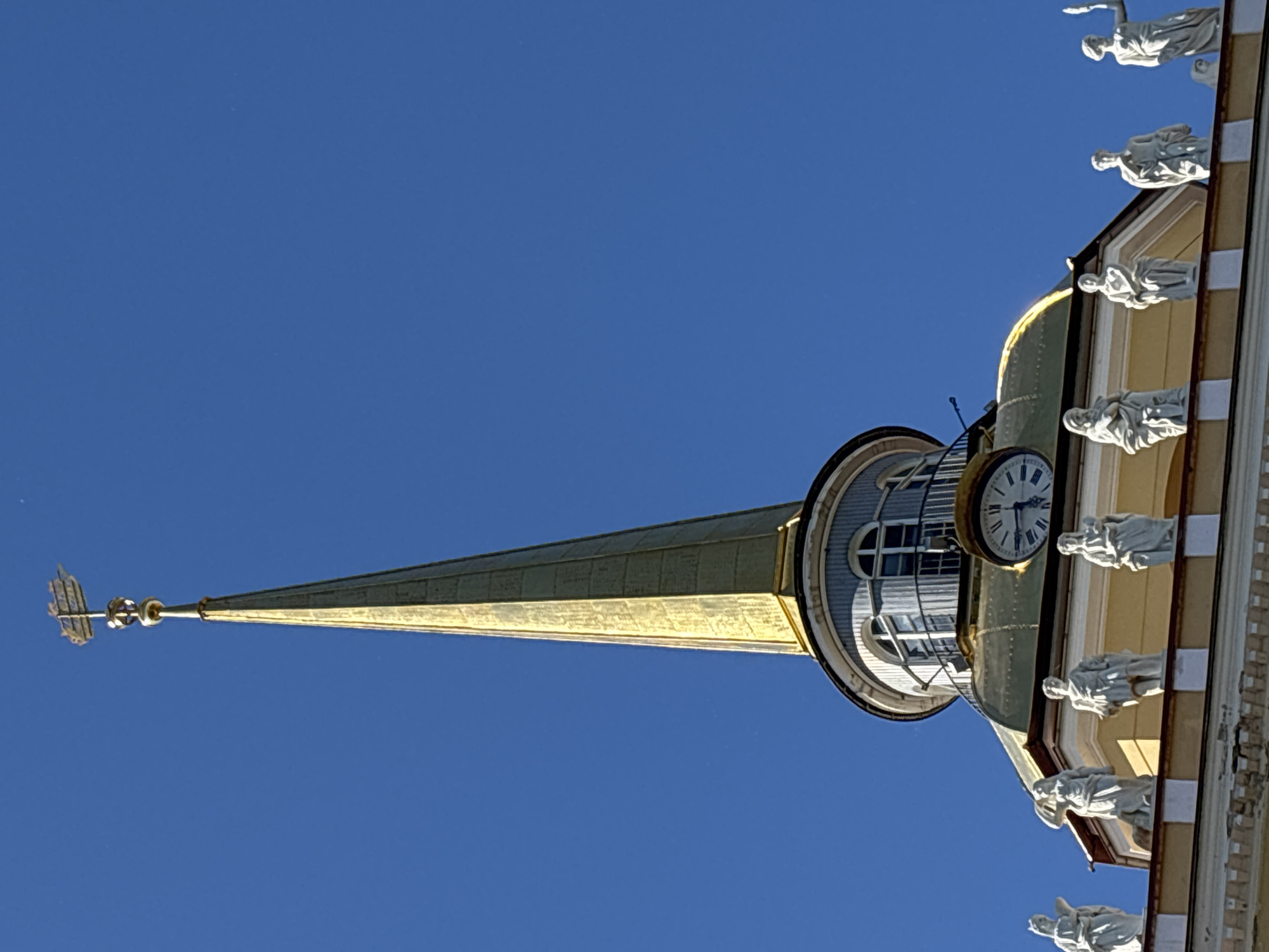This is a low-angle shot of a church spire against a clear blue sky. The spire is golden and tapers to a point, topped with a decorative finial. Attached to the side of the spire's base is a clock tower with a white face and black Roman numerals. The clock appears to be stopped at approximately 1:57. The building has a yellow facade with ornate white trim and is adorned with several white statues of figures in flowing robes, positioned as if overlooking the scene below. These statues appear to be in niches or protrusions from the building. The sunlight suggests a bright, clear day. The presence of classical architecture and the spire's design are indicative of a European, specifically Russian, location like Saint Petersburg, as suggested by the provided context. No visible text is present in the image.