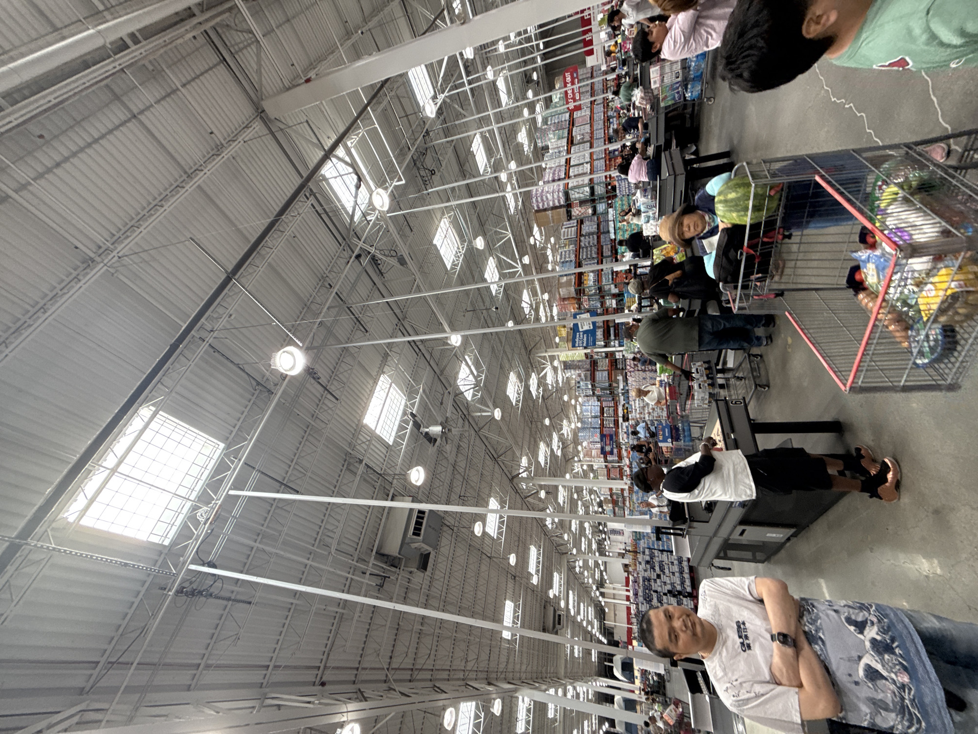 The image depicts the interior of a large warehouse-style retail store, likely a Costco, located in the City of Yonkers, United States of America. The scene is populated with numerous shoppers and employees. The architecture features a high, corrugated metal ceiling with exposed beams and numerous fluorescent lights and skylights. Racks of merchandise, including boxes of food and household items, are visible throughout the store.
In the foreground, a man wearing a white graphic t-shirt and jeans stands with his arms crossed, looking towards the camera. To his right, another man in a white and black t-shirt and black shorts stands at a checkout counter. In the middle ground, several people are pushing shopping carts and interacting at checkout stations. One woman in the middle ground is wearing a wide-brimmed hat and looking at a watermelon in her shopping cart. Further back, more shoppers are visible browsing merchandise and moving through the store. The overall atmosphere appears to be one of a busy shopping day.