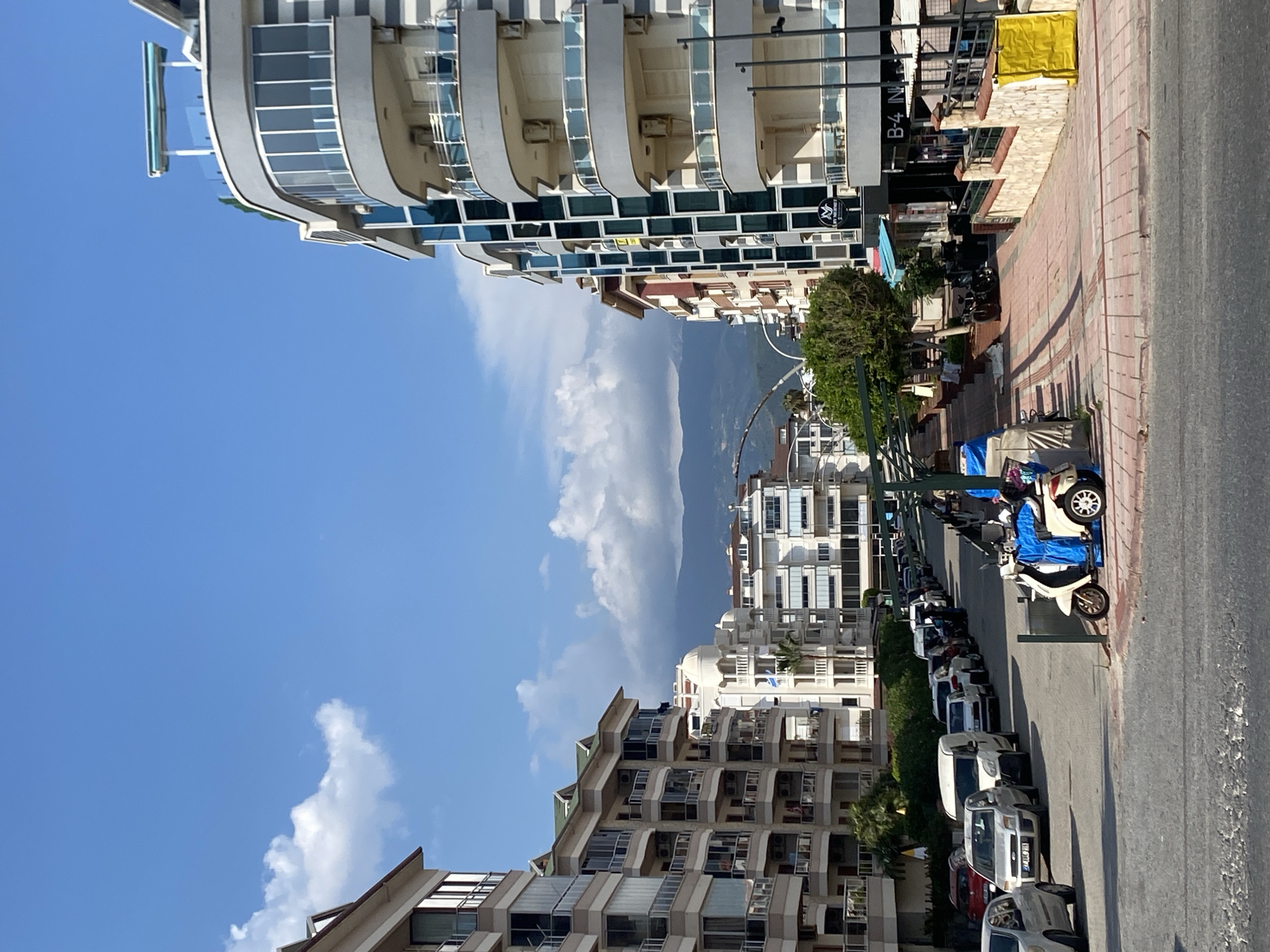 This image captures a street scene in Alanya, Turkey, likely in a residential or commercial area with apartment buildings. The street is lined with multi-story buildings, featuring balconies and modern architectural elements. Several cars are parked along the street on the left side, and a scooter is visible on the right. The sky is a bright blue with scattered white clouds, and mountains are visible in the distance. The street itself appears to be paved, with a pedestrian walkway lined with trees and shops.