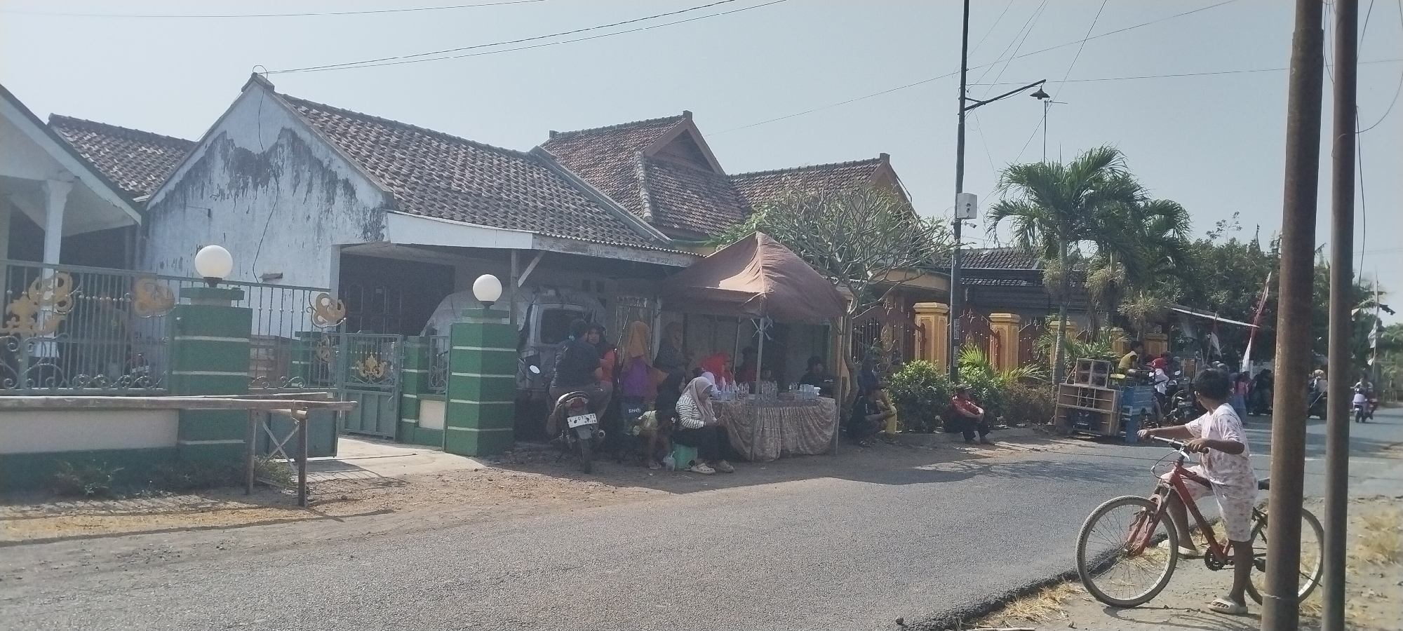 The image depicts a street scene in Yosowilangun Kidul, Indonesia, likely in front of a residential area. A row of houses with tiled roofs lines the street. In the foreground, a child wearing patterned shorts and a t-shirt stands next to a red bicycle. A paved road occupies the lower portion of the image. To the left, a white house with a green and white fence and gate is visible. Further down the street, several people are gathered around a stall with a brown canopy, possibly a market or gathering. A motorcycle is parked nearby. Palm trees and other vegetation are present. The sky is clear and bright, indicating daytime. The overall impression is that of a typical village or small town street with everyday activity.