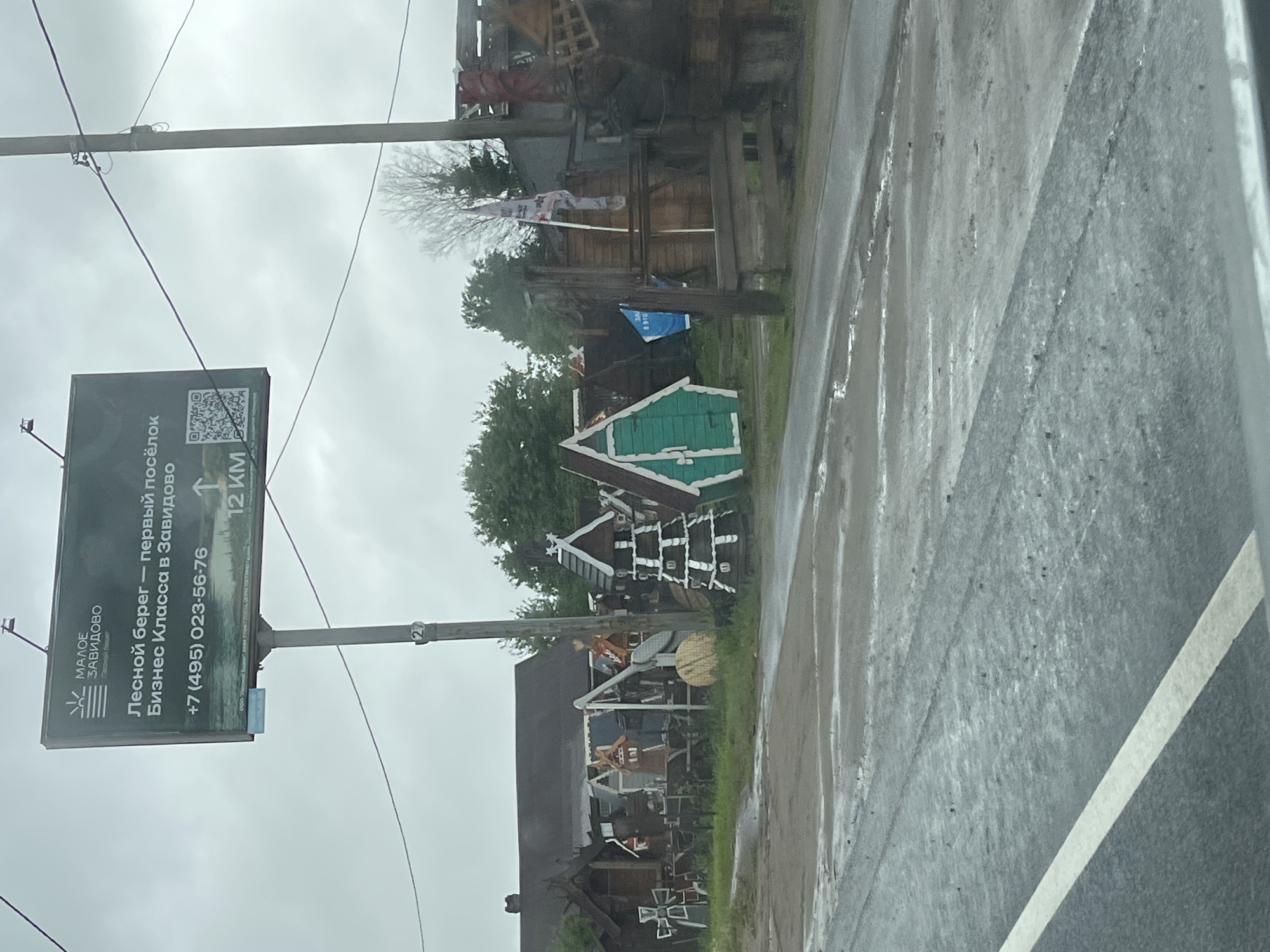 The image depicts a street scene in Zavidovo, Russia, likely taken on a rainy day based on the wet asphalt. A large advertising billboard is visible to the left, promoting a business class real estate venture called "Forest Shore." The sign displays the phone number +7 (495) 023-56-76 and indicates the location is 12 km ahead. In the background, a cluster of small, ornamented wooden structures is present on the roadside, possibly decorative or part of a local attraction. Overhead utility lines are visible. A partial view of a vehicle is seen in the lower right corner of the image.