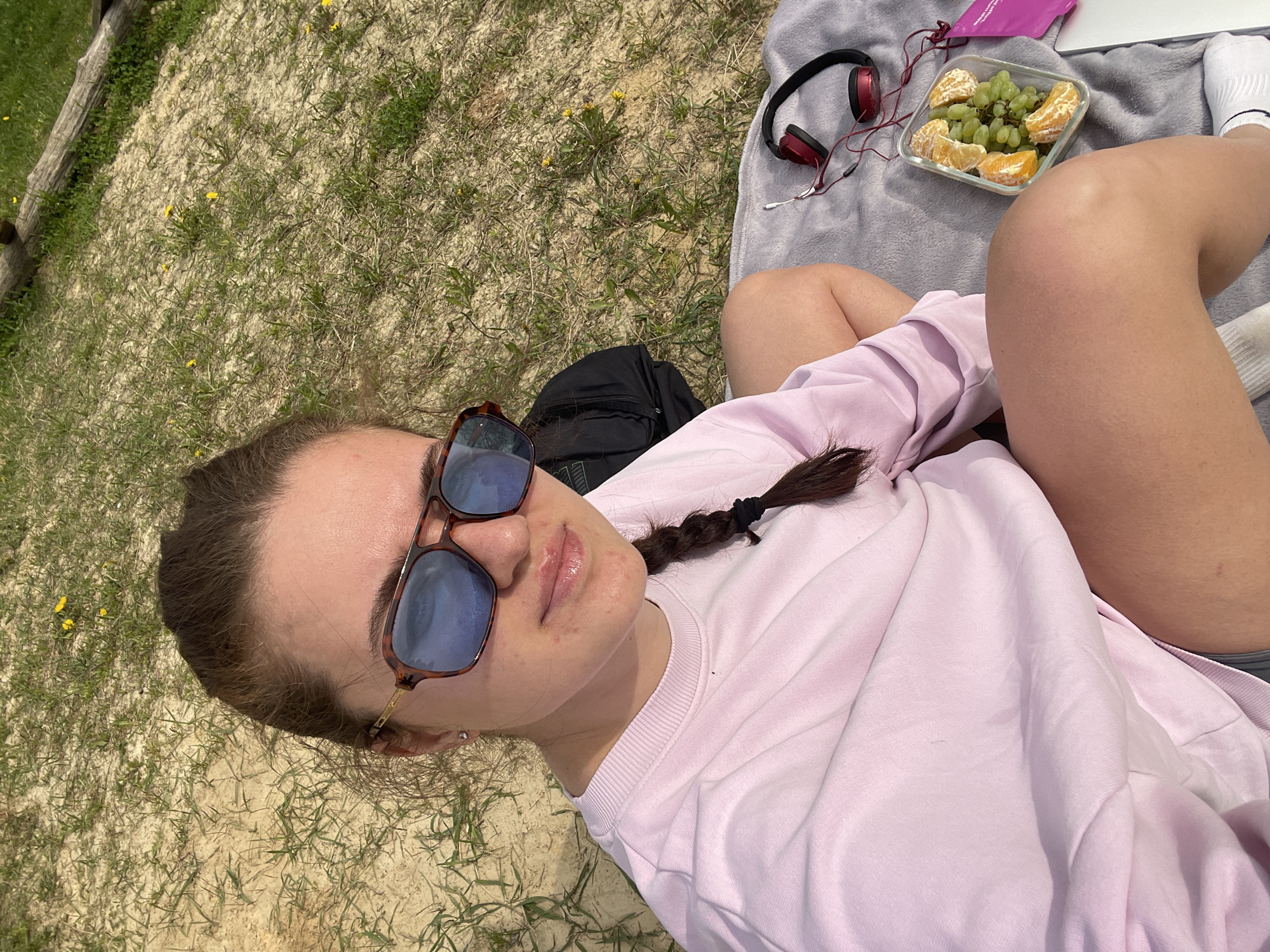 A young woman is reclining outdoors on a patch of mixed grass and dry ground. She is wearing sunglasses and a light pink long-sleeved shirt, with a dark braid. To her side is a gray blanket with headphones and a container of grapes and orange slices. A backpack is nearby, along with what appears to be a laptop and a pink packet. The location is unknown.