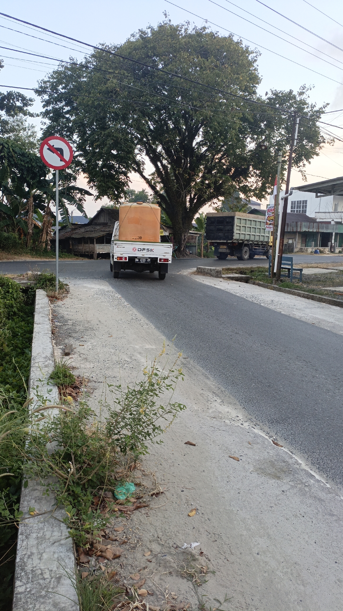 In Gusunge, Indonesia, a white pickup truck with a large orange water tank on its bed is captured in the middle of a paved road. The truck, branded with "DFSK," bears the license plate "DA 9703 ZP." It is positioned to the left of a larger, dark-colored dump truck, suggesting a traffic scenario. The road is flanked by a concrete bridge on the left, with dense green foliage growing on its embankment. On the right side of the road, a bench sits beside a building with white siding and multiple windows, possibly a shop or residence. A large tree with dense green foliage dominates the background, its branches reaching towards a clear sky. In the foreground, to the left of the road, a red and white traffic sign prohibiting left turns is visible, indicating a specific traffic regulation for this area. The overall scene depicts a typical street view in an Indonesian city, with vehicles, infrastructure, and natural elements coexisting.